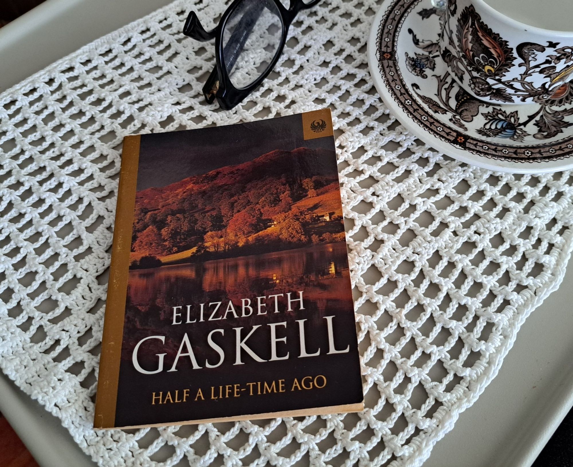 The book Half a time ago by Elizabeth Gaskell, short story with an autumnal colored cover with a tea cup, glasses over a crochet cloth