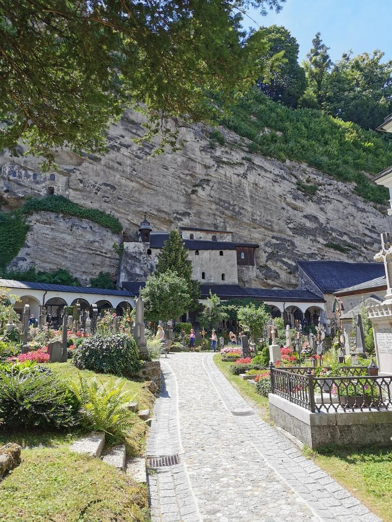 Am felsigen Mönchsberg der alte St Peter-Friedhof. Im Vordergrund kleinere Gräber. Nah am Berg ein grauer Bau und Grabstätten. Der Fels grau mit Grün bewachsen. 
