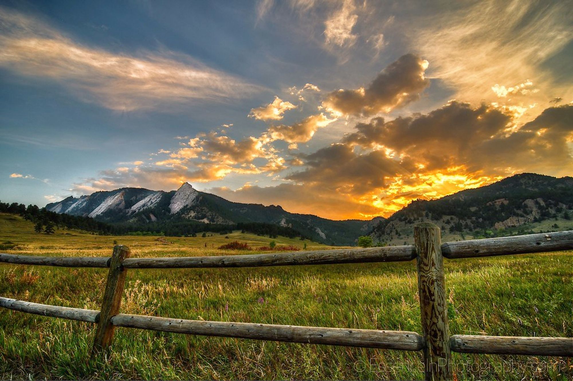 photograph of the Flatirons mountains in Boulder, Colorado at sunset. dark blue sky with clouds radiating across the sky. the sun has just set behind the mountains. a meadow of green grass beneath the mountains with a wood fence in the foreground.