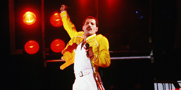 Icónica fotografía de Freddie Mercury con la ropa blanca y la chaqueta amarilla que le caracterizó durante el tour Magic, levantando el brazo en su gesto más característico sobre el escenario. Discogs.