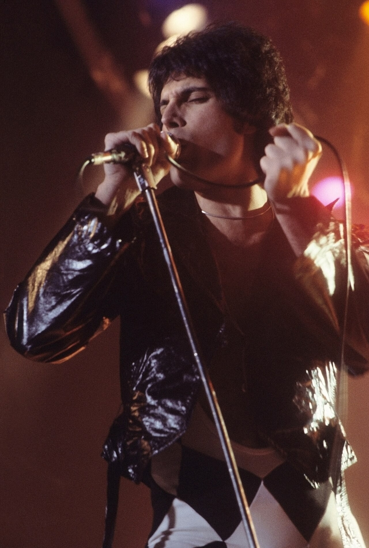 Freddie Mercury con unas mallas en rombos negros y blancos y una chaqueta de cuero a finales de los 70, sobre el escenario. Fotografía de Carl Lender • CC BY-SA 3.0