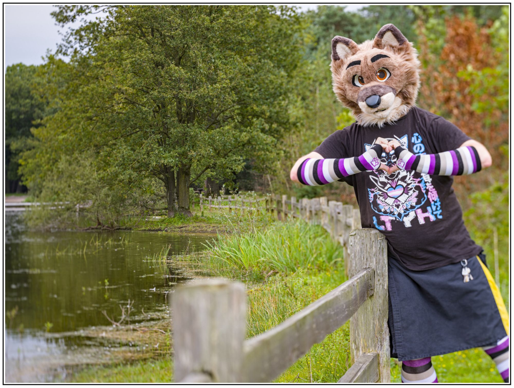 A wolf fursuiter in a kilt wearing ace flag colored arm warmers, they are in a forest the wolf looks to be photographed with a shallow depth of field even though a tree far in the background is also sharp! 