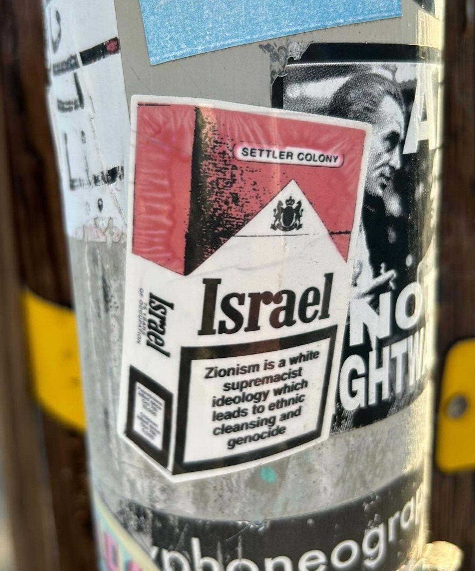 Sticker on a metal pole, made to look like a 3 dimensional pack of Malboro cigarettes. 

The upper pack is in red, with the words "settler colony" inside it. Underneath is a coat of arms and then "Israel" in large, black on white text.

The bottom of the pack has a black bordered square where cancer warnings go. Inside the square, it says "Zionism is a white supremacist ideology which leads to ethnic cleansing and genocide"