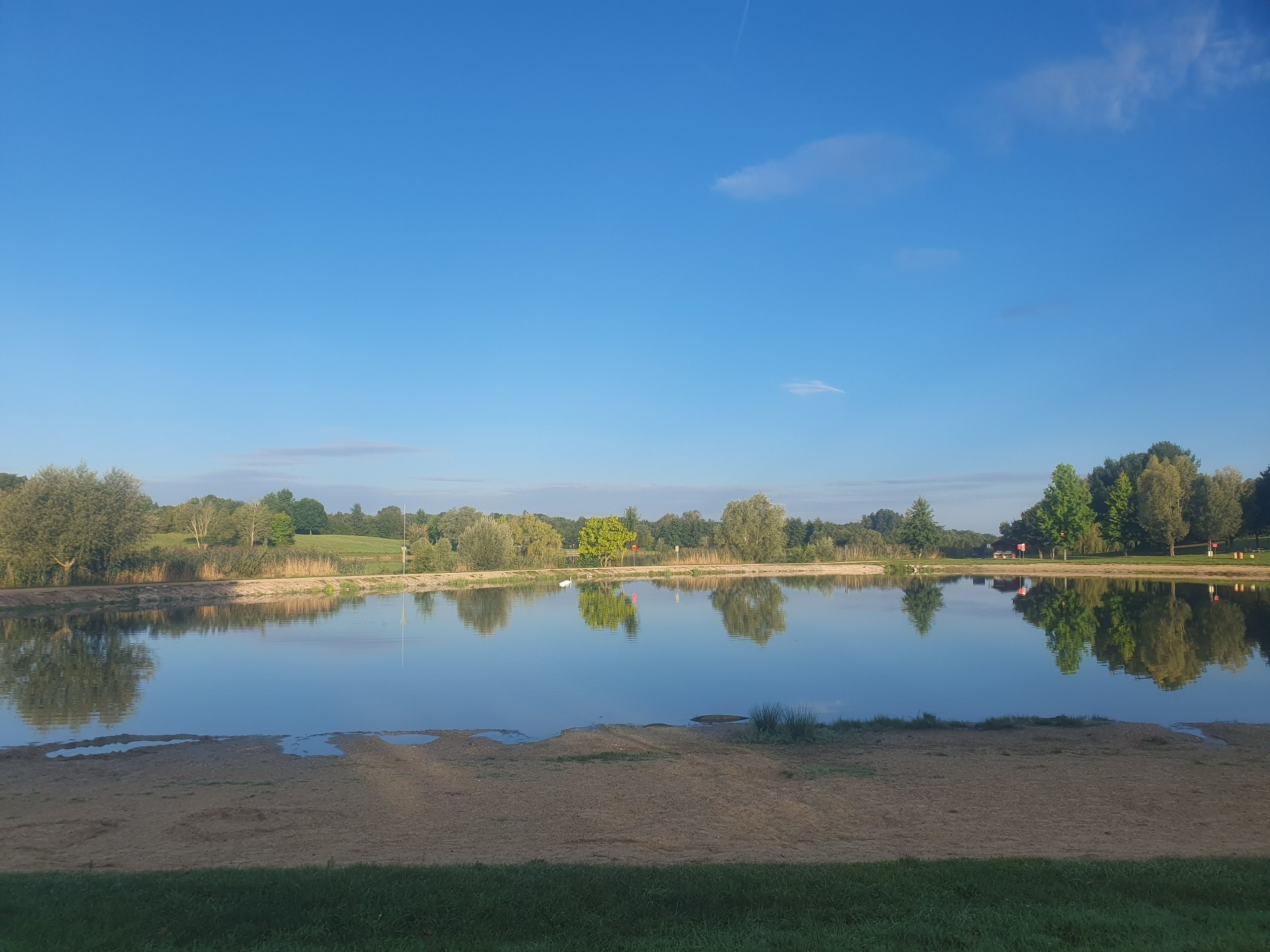 Ein kleiner See im Morgenlicht.
Im Vordergrund ein kleiner Strand. Am anderen Ufer Bäume. Blauer Himmel.