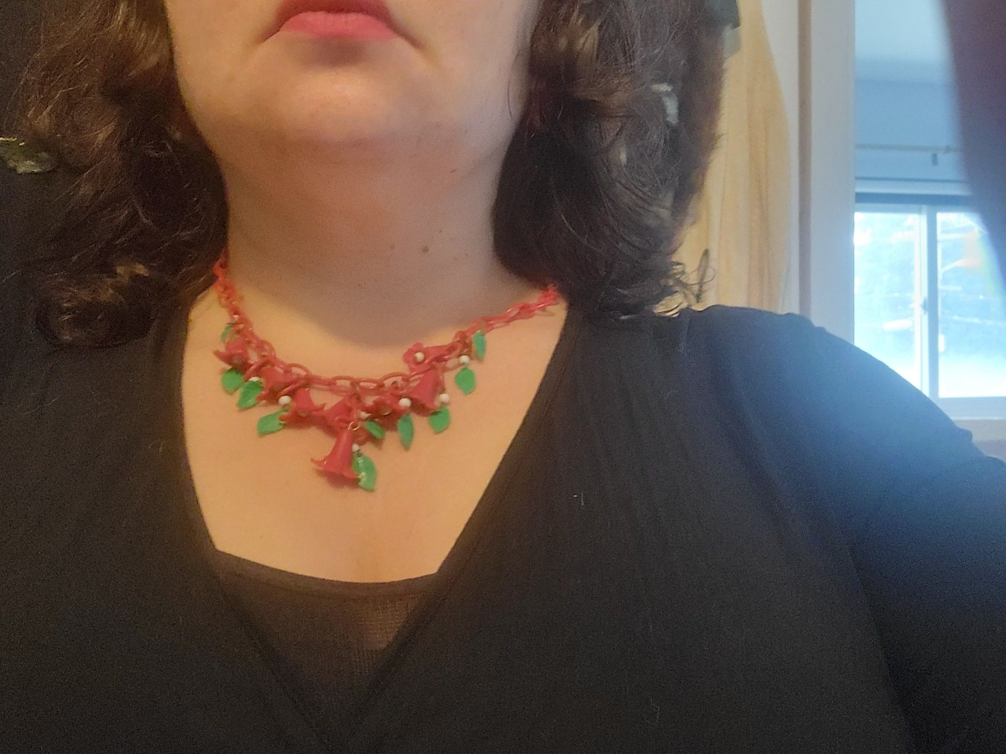 My light skinned torso in a black wrap top. Visible on my neck is a red celluloid chain necklace with red celluloid trumpet flowers pointing down. There are white beads in the middle of the trumpet and green leaves hanging next to them. You can also see my down turned mouth (I have RBF) wearing red lipstick, and my square-ish jaw.