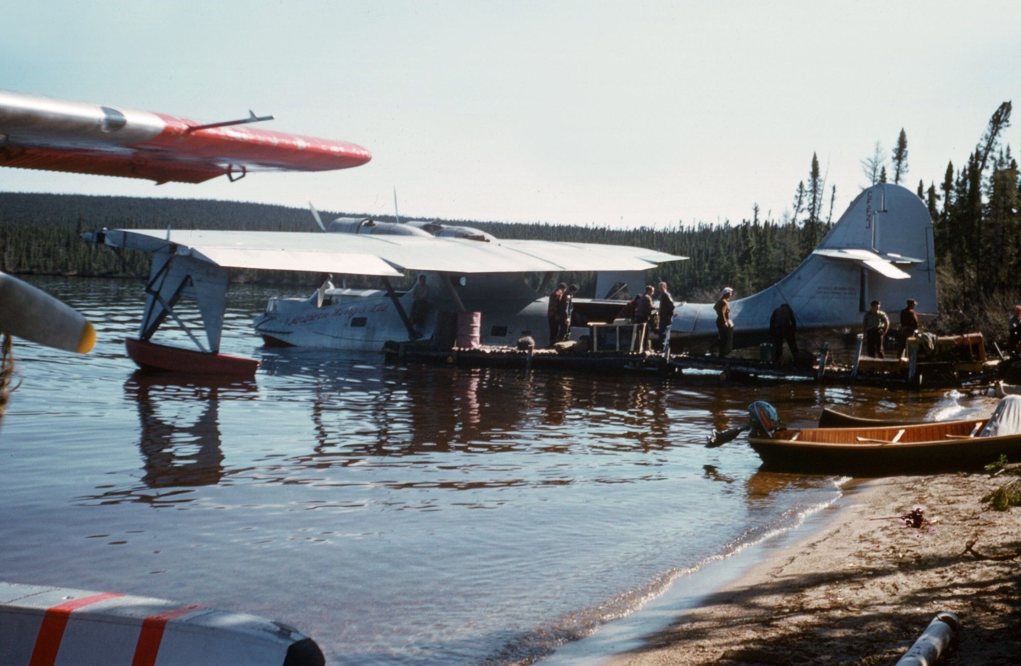 An image of a beach on a lake with float planes being loaded with supplies, and small boat pulled up onto the sand.