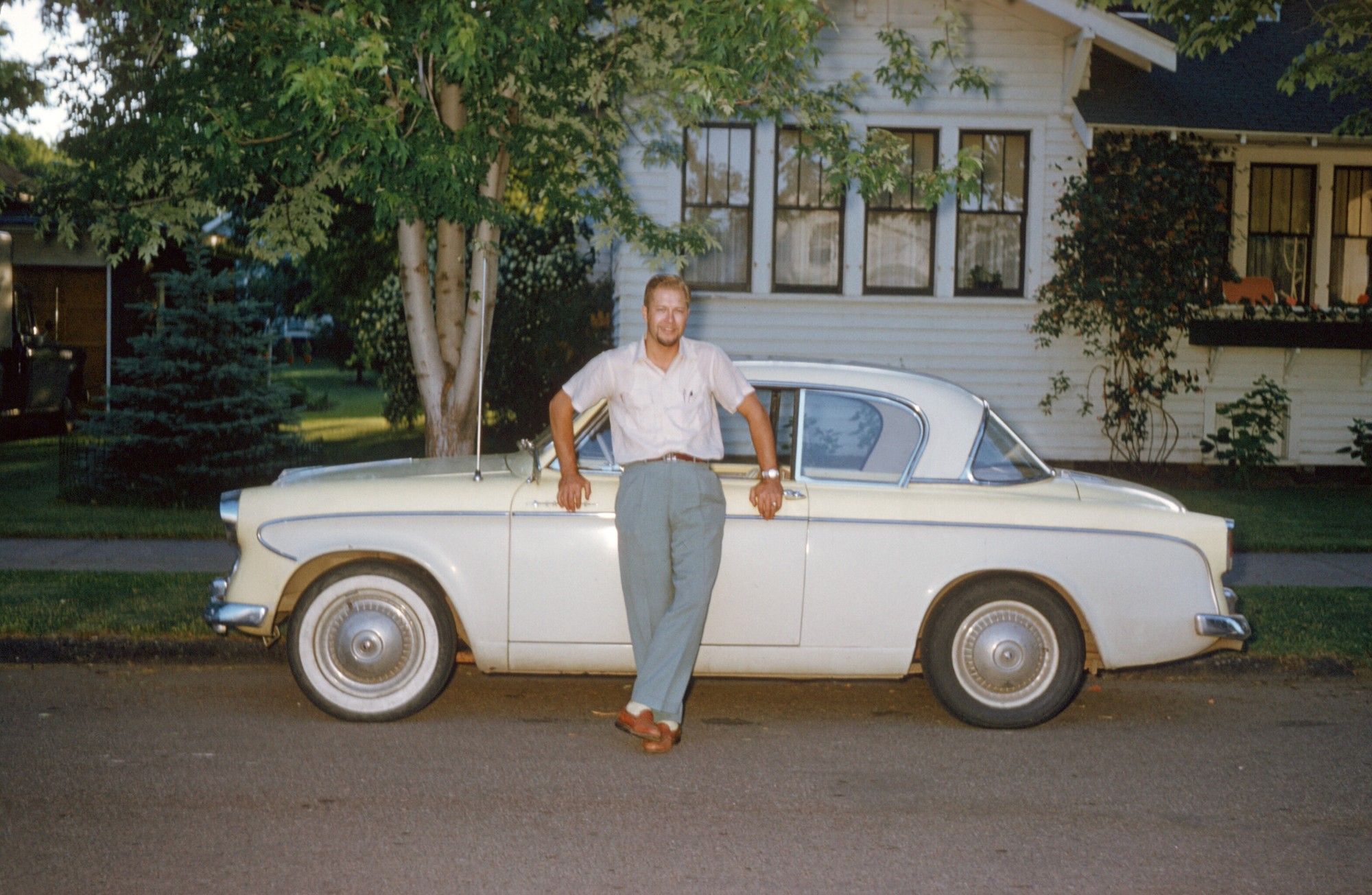 A photograph from the 1950s, exterior, picture of a young man in slacks and short-sleeved shirt standing in front of a small, sporty car in grey and white. The street is obviously a residential street as you can see trees and a house behind the car.