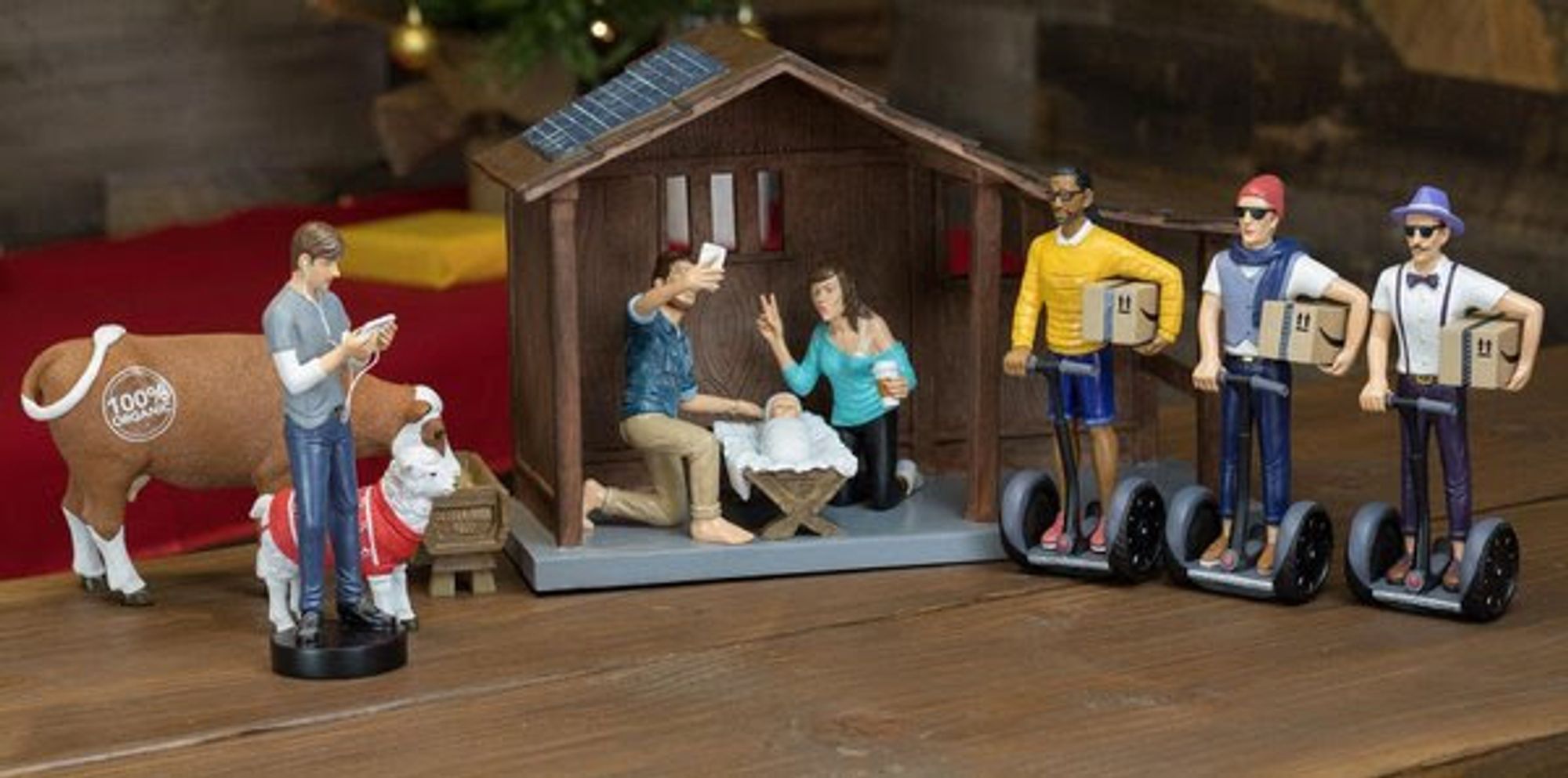 A version of a creche but the three wise men are on Segway scooters, dressed in Hipster outfits and carrying Amazon boxes. Mary and Joseph in modern clothes are taking a selfie with the Baby Jesus and the shepherd is checking his phone next to a sheep in a sweater, and a cow with a "100% organic" sticker on it.