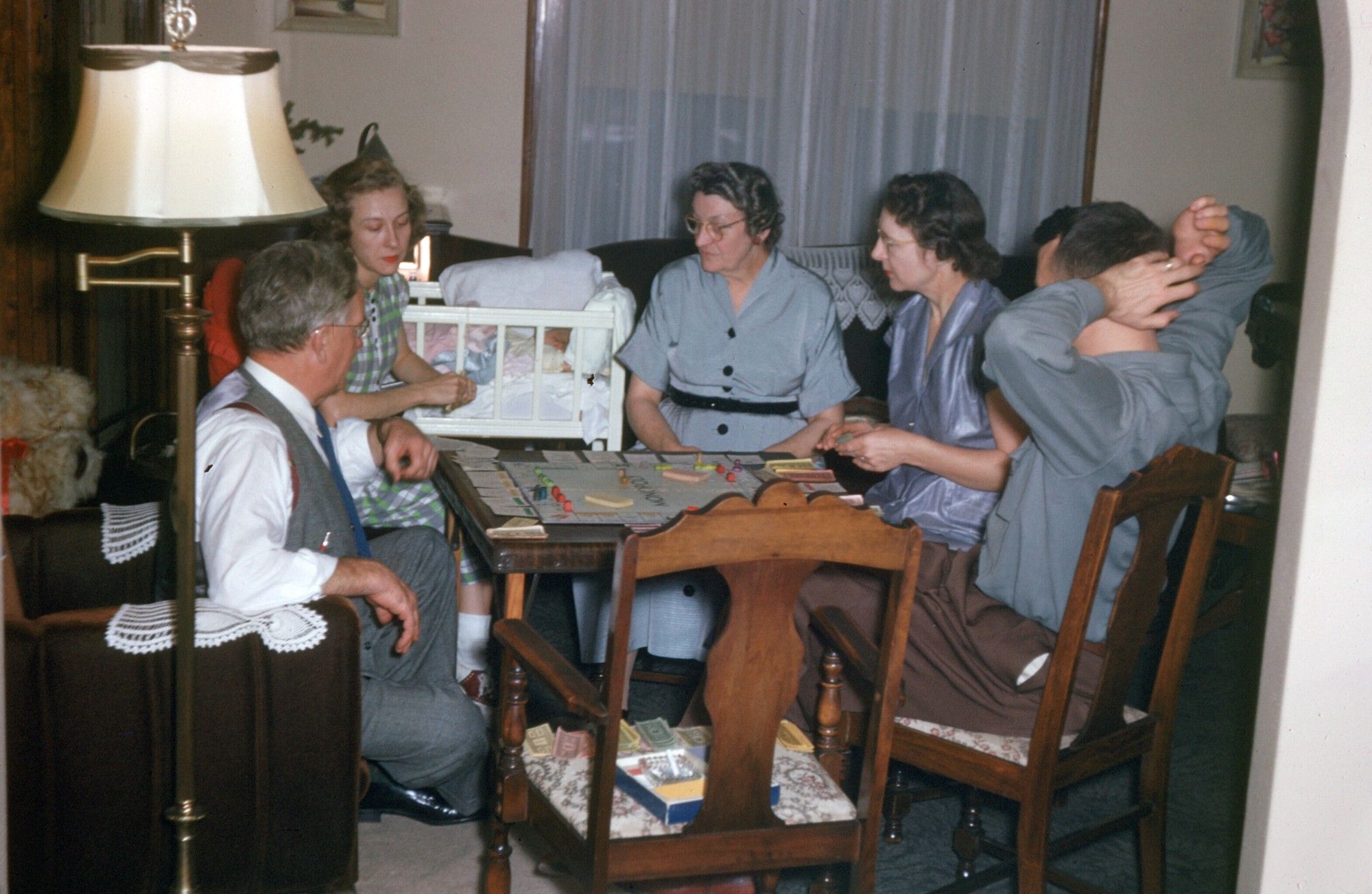 A photograph from the 1950s where you have three women and two men sitting around a card table playing Monopoly. There is a baby bassinet in the background and an empty chair for the photographer in the foreground.