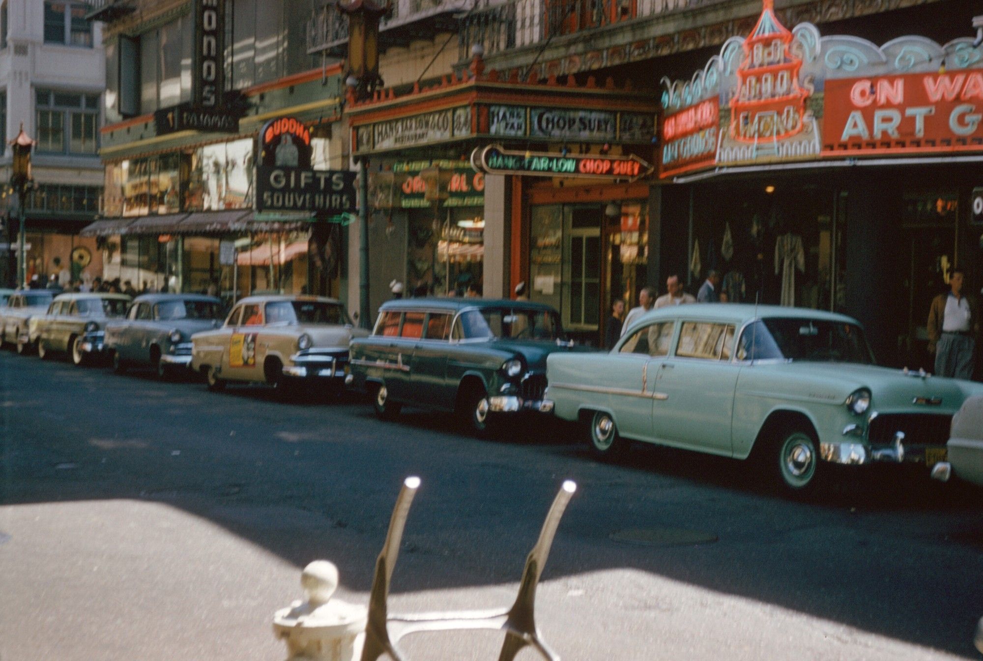 1950s Chinatown in San Francisco. A street scene with cars parked along the street in front of gift and souvenir shops with gaudy and even neon-lit signs.