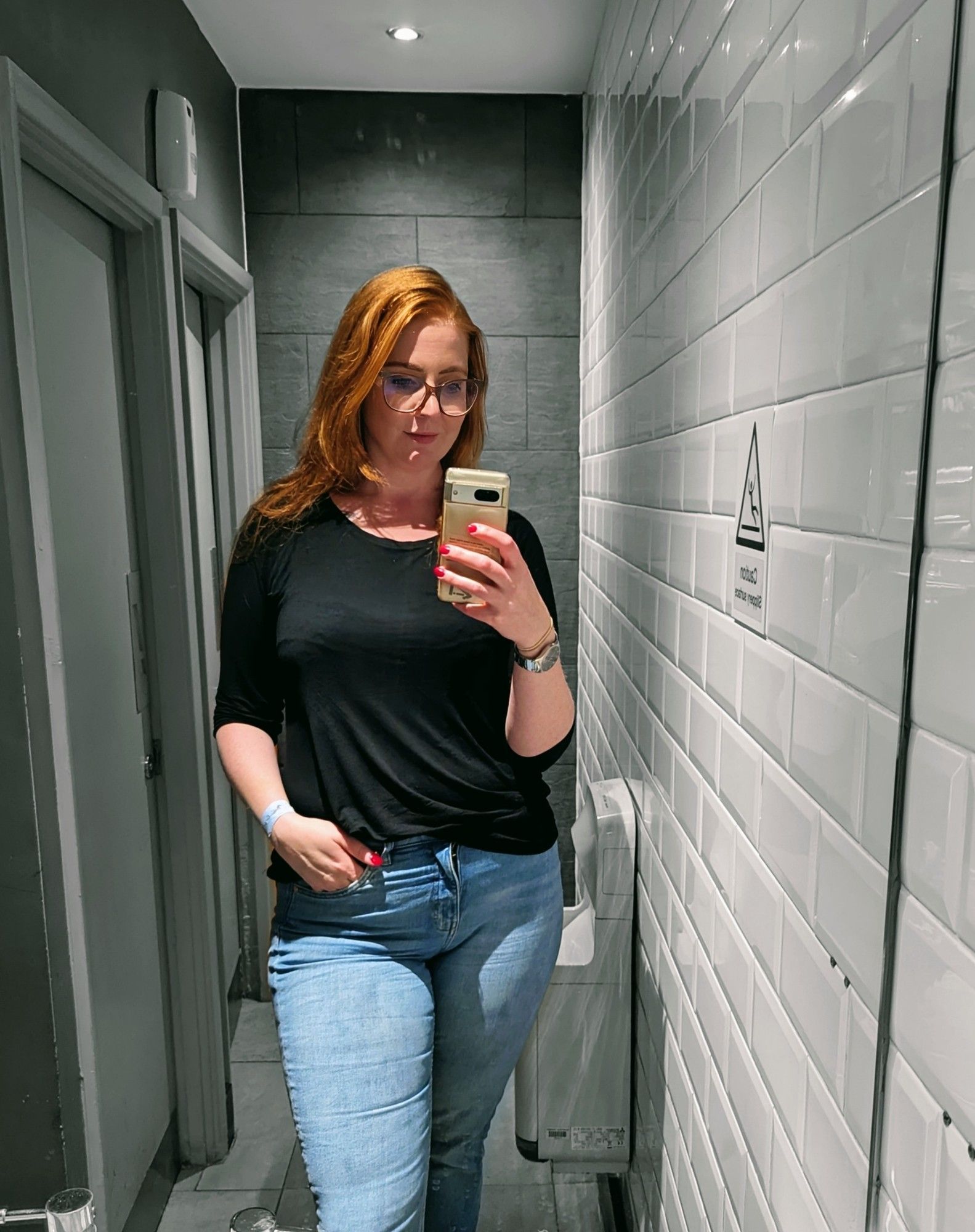 Spegelselfie på mig, jag har blåa jeans, svart topp, utsläppt rött hår, ena handen i fickan och småler på en toalett tillhörandes en resturang