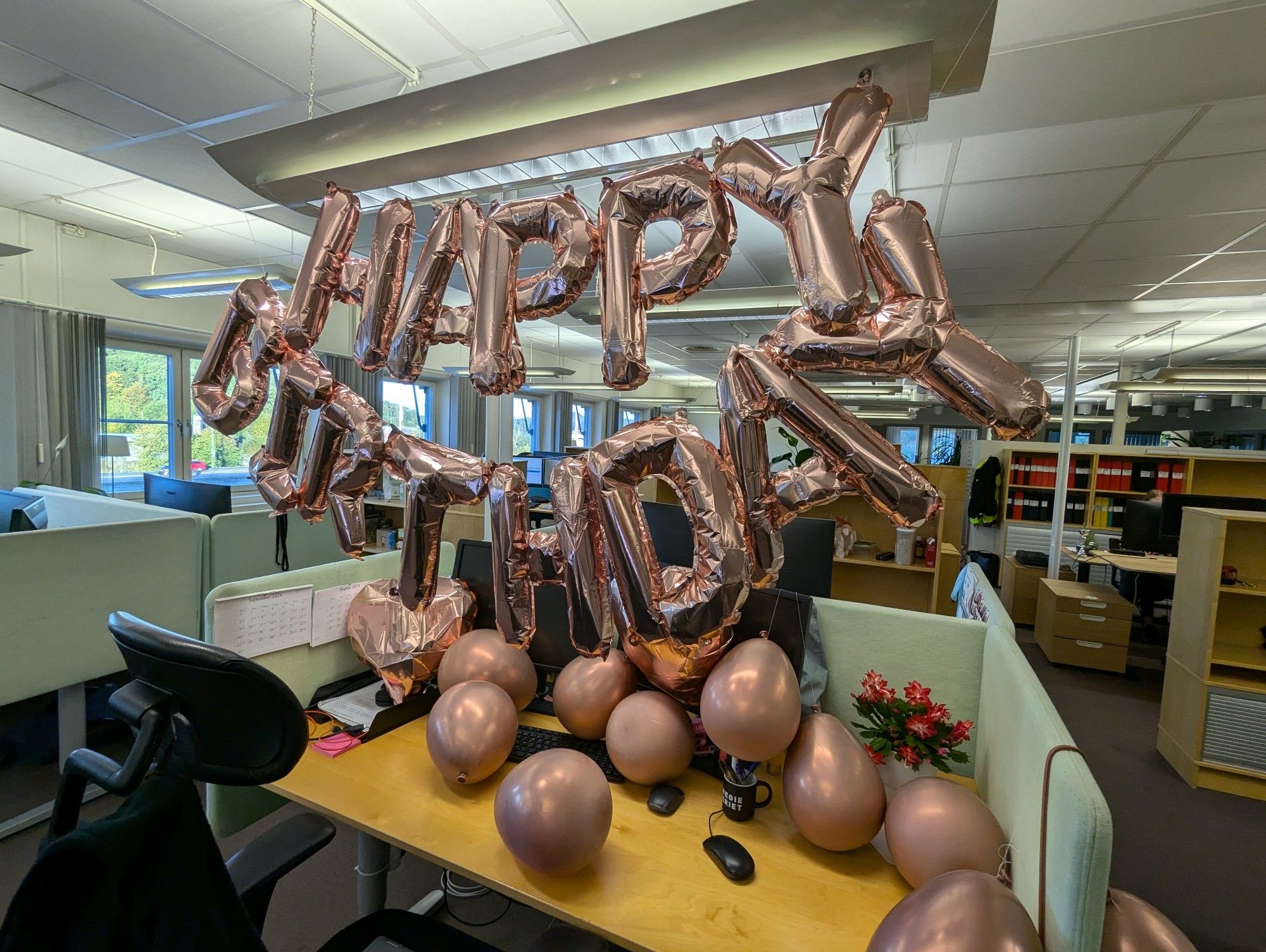 Skrivbord i öppet kontorslandskap. På skrivbordet ligger flertalet rosaguldiga ballonger. I lampan ovanför skrivbordet hänger ballonger som bokstaverar Happy Birthday