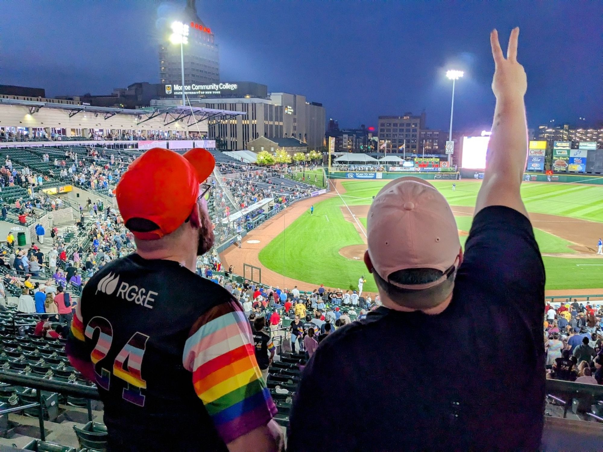 Two figures looking out of the suite balcony towards the baseball field, one of them holding up two fingers.