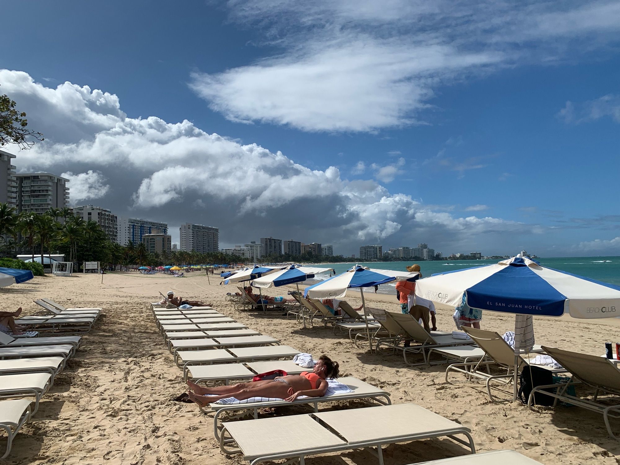 Photograph taken down a Puerto Rican beach. The water is somewhat blocked by beach umbrellas but what can be seen is blue/green. There are many beach chairs laid out, with a few people scattered between and on them. Down the beach are condos and hotels. Above them, rain clouds can be seen moving towards the ocean.