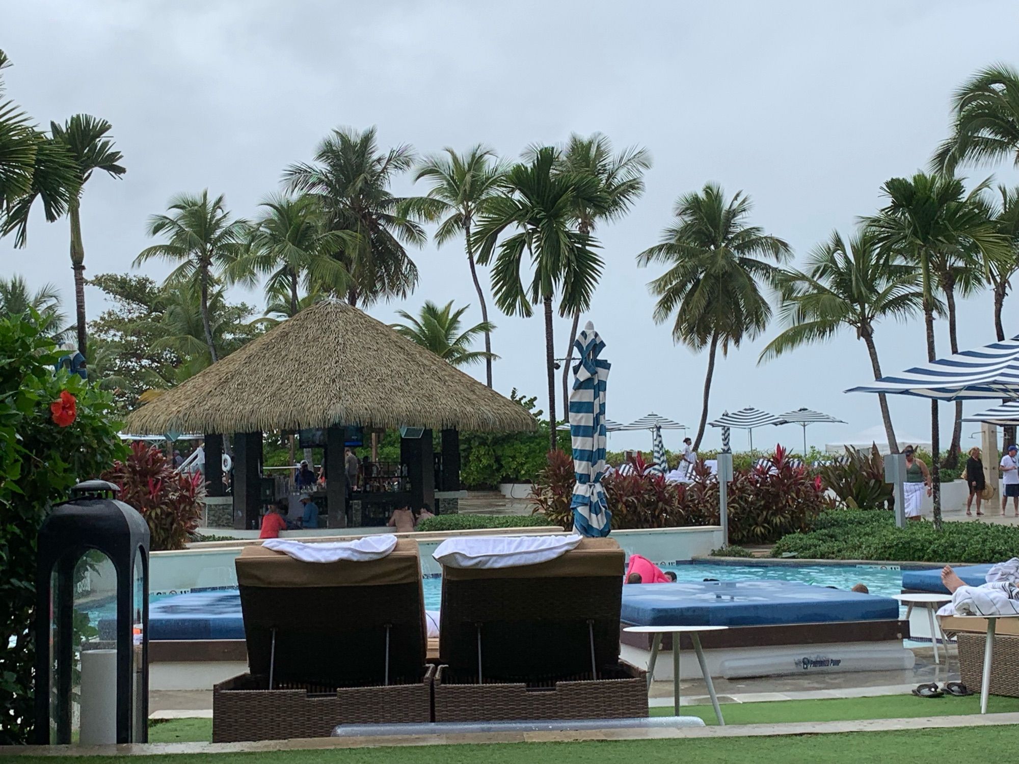 Pool view from a resort cabana. There are blue lounge beds and two propped up loungers facing away from the camera. A swim up tiki bar can be seen across the pool. There are many palm teens around the pool and bar