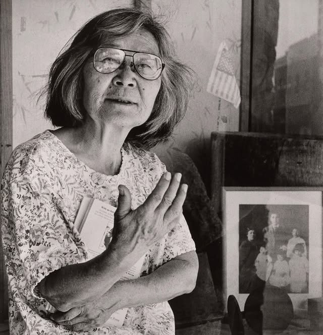 Writer Hisaye Yamamoto circa 1980s. She is holding a book to her chest and standing next to a small family portrait.