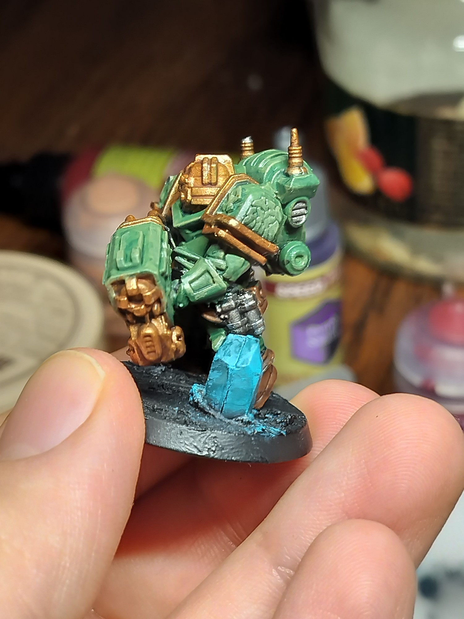 A deep rock galactic dwarf with green armour with pale green highlights and gold trim. A blue crystal is on the base 