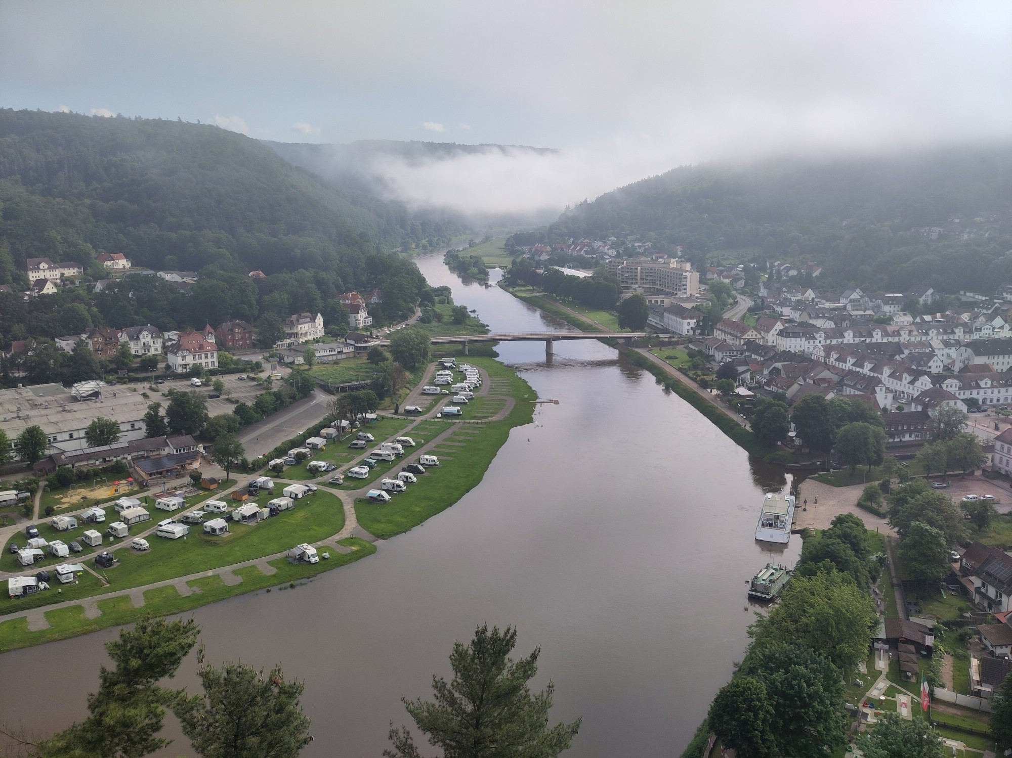 Ein Fluss schlängelt sich durch bewaldete Bergen man sieht einen Campingplatz und eine Stadt am Ufer. Es gibt eine Brücke. Aufnahme von oben.