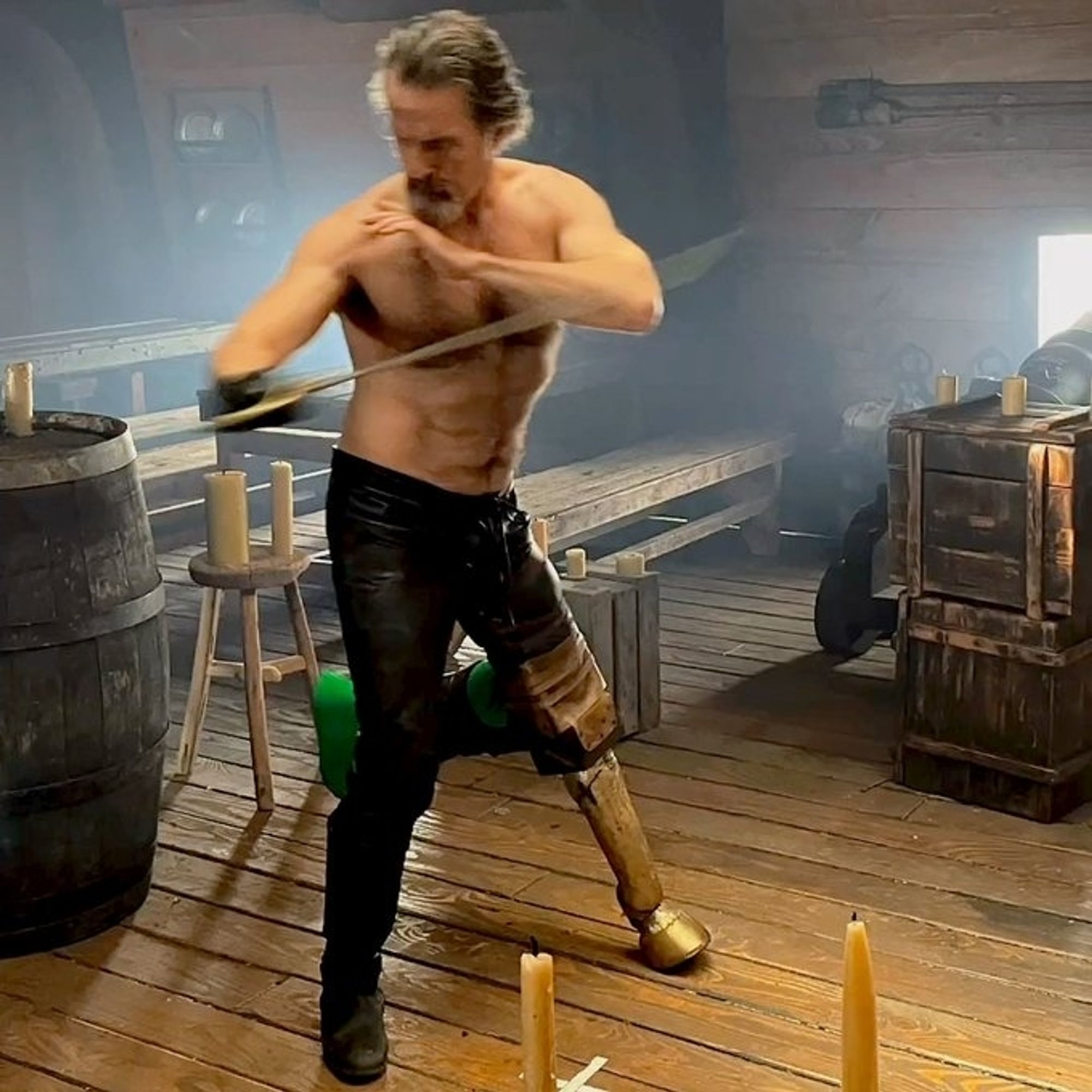 Screenshot of behind the scenes footage of Con O'Neill in season 2 episode 5 of Our Flag Means Death, the shirtless swordplay scene. His hair is pushed back but loose, blurred in motion as he swings the sword at pec height. Muscles, furry chest, tight leather pants. 

He is wearing the wooden leg prosthetic, with his real leg in a green screen sock behind him and supported at 90 degree angle.