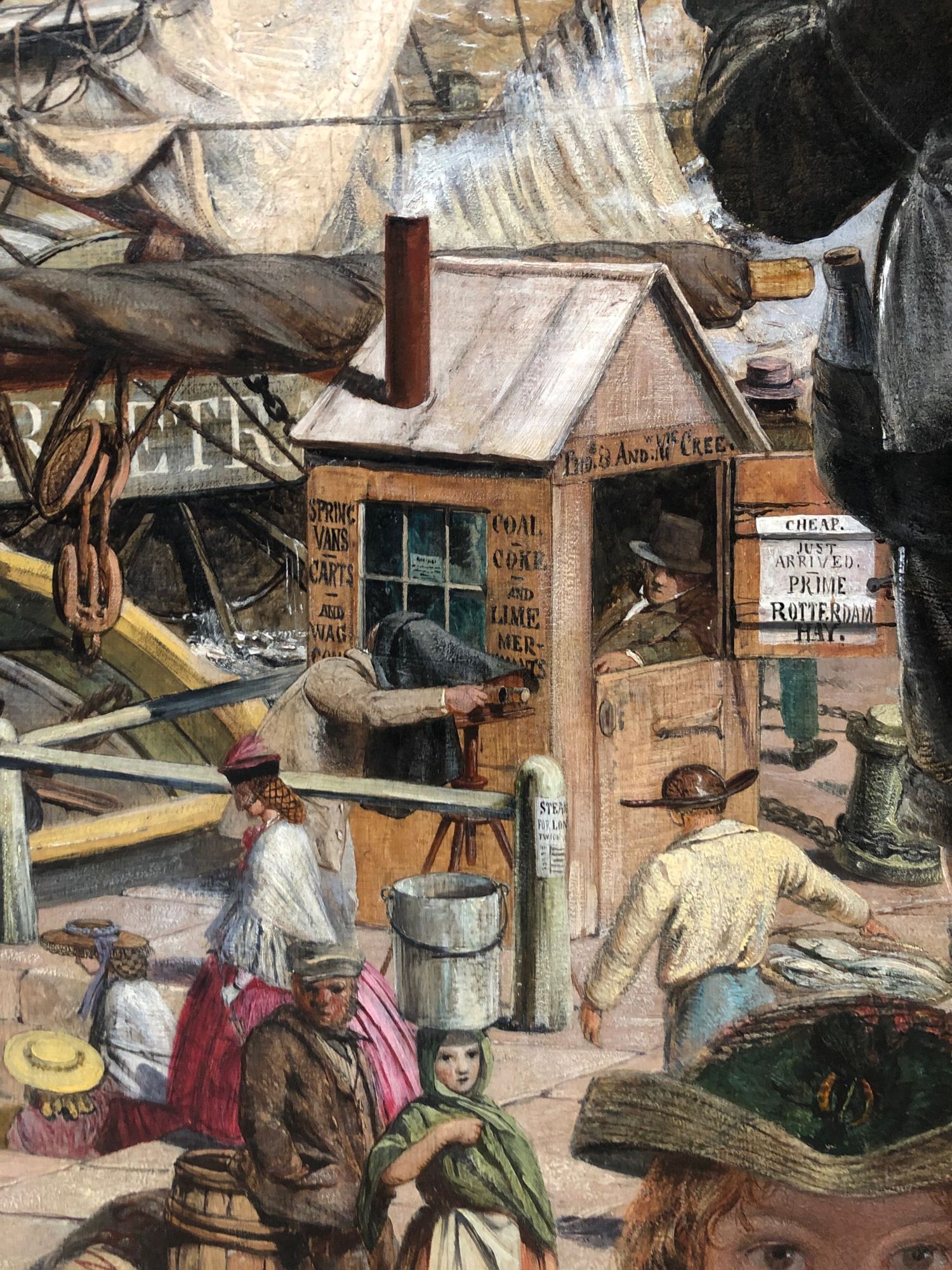 Detail of an oil painting showing activity on the banks of the Tyne at Newcastle in 1861: rolled sail on a ship beam at top, a wooden hut with a man seated inside, advertising cart transport, coal and coke, and Rotterdam hay, and a mix of figures at work or rest - including a photographer under the dark cloth of a sterescopic camera. By William Bell Scott, at Wallington house, National Trust, near Morpeth, Northumberland.