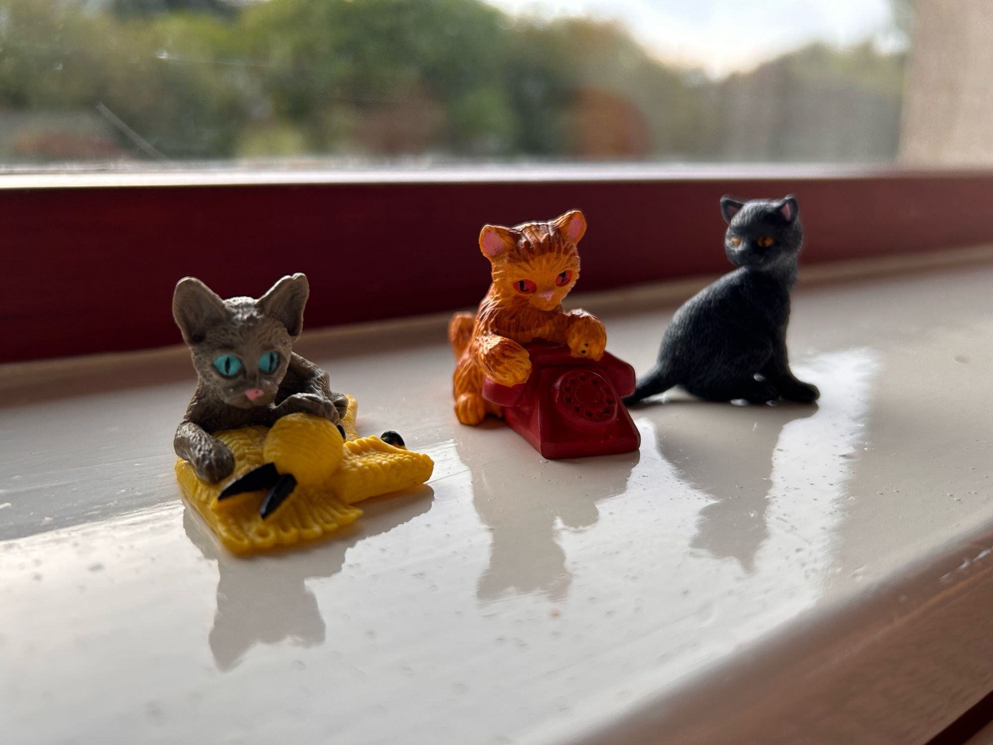 Three miniature toy cats. One is brown and sitting on knitting. The next is ginger and sitting by a red phone. The last is black.