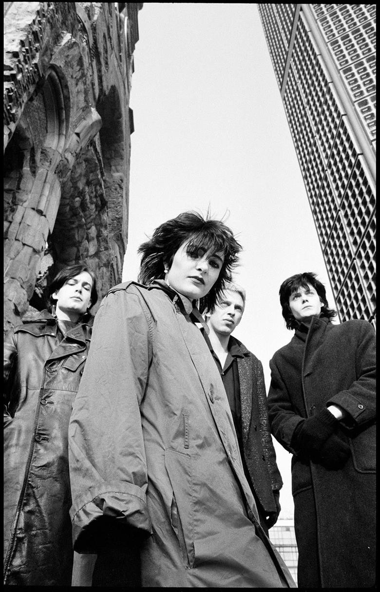 Foto dal basso ai Siouxsie and the Banshes di fronte a una struttura architettonica e un palazzo molto alto. Sulla sinistra c'è un antico edificio in pietra con una finestra ad arco, la cui superficie è strutturata in pietra. Sullo sfondo a destra si erge un grande edificio moderno, caratterizzato da una facciata a griglia con numerose finestre identiche. 