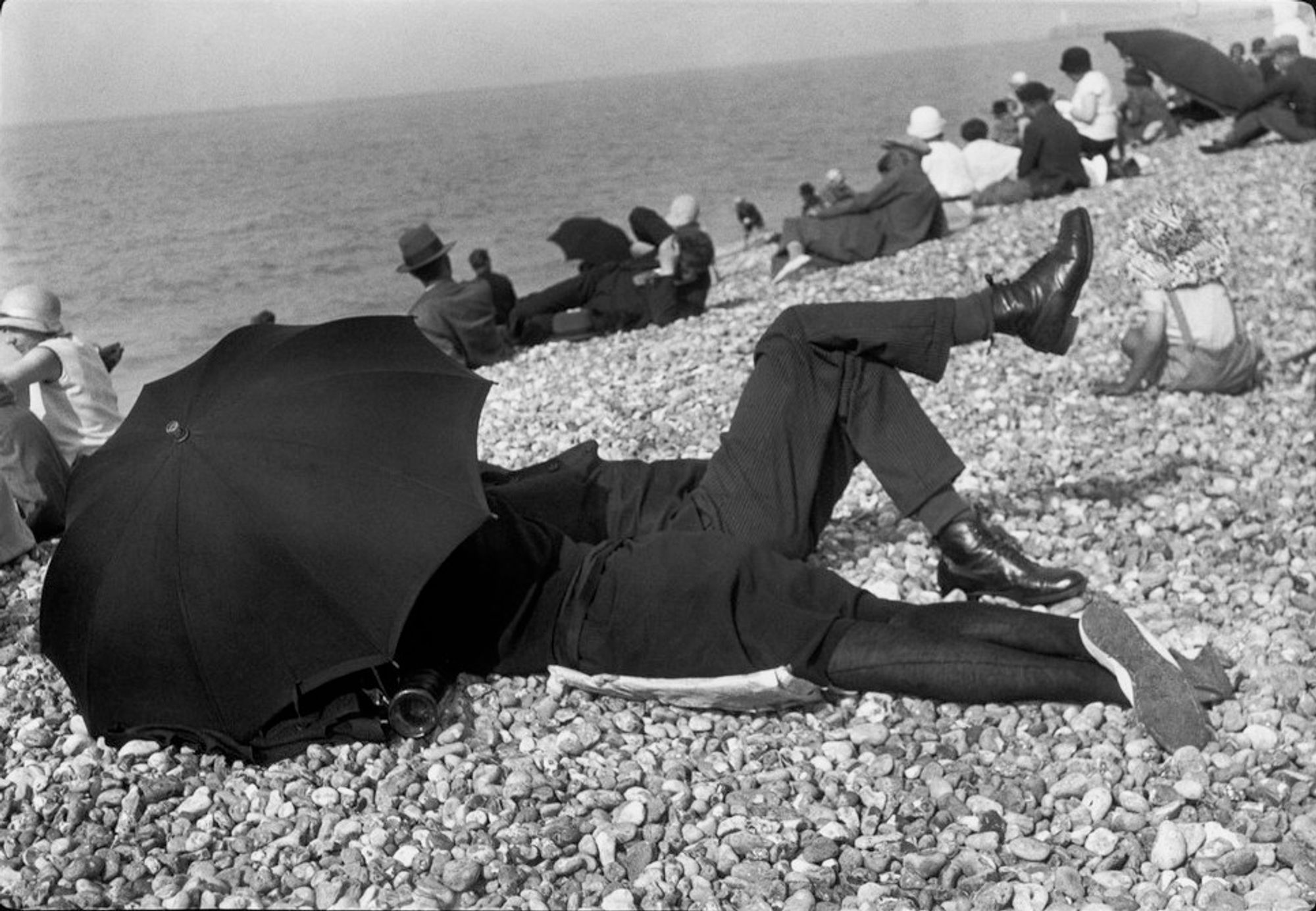 Una foto in bianco e nero fatta in una spiaggia di sassi, dove diverse persone si rilassano. In primo piano, un uomo e una donna sono sdraiati sulle pietre, vestito lui con un abito scuro e scarpe lucide e lei con vestito corto e calze scure. Sono sotto un grande ombrello nero che gli copre il viso. Sono visibili solo le gambe delle persone e parte delle scarpe. Accanto a loro una bambina piccola seduta. Dietro di loro, altri bagnanti sono seduti o sdraiati, alcuni in giacca e cravatta, altri in abiti casual. Lo sfondo mostra un'ampia vista del mare sotto un cielo limpido. 