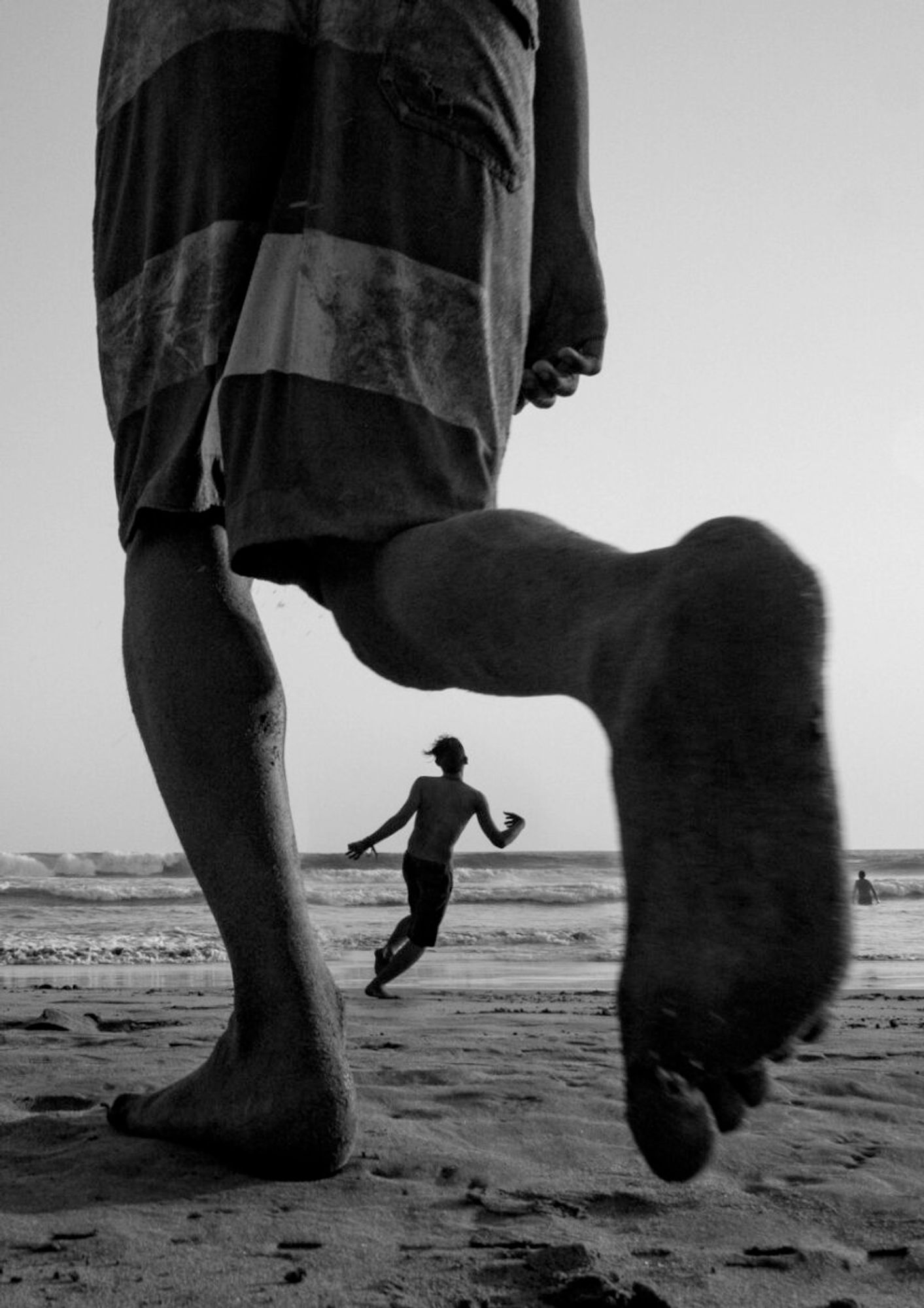 L'immagine è una scena dinamica della spiaggia in bianco e nero. In primo piano, la metà inferiore di una persona viene catturata a metà passo, sottolineando un piede e una gamba che avanzano sulla spiaggia sabbiosa. La gamba e il piede occupano una parte significativa dell'inquadratura, fornendo un senso di scala e movimento. Questa persona indossa pantaloncini a righe. Nella via di mezzo, un'altra persona, a torso nudo e in pantaloncini, corre verso il mare, con il corpo inclinato in movimento. Lo sfondo mostra le onde del mare che si infrangono sulla riva, sotto un cielo limpido senza nuvole visibili.