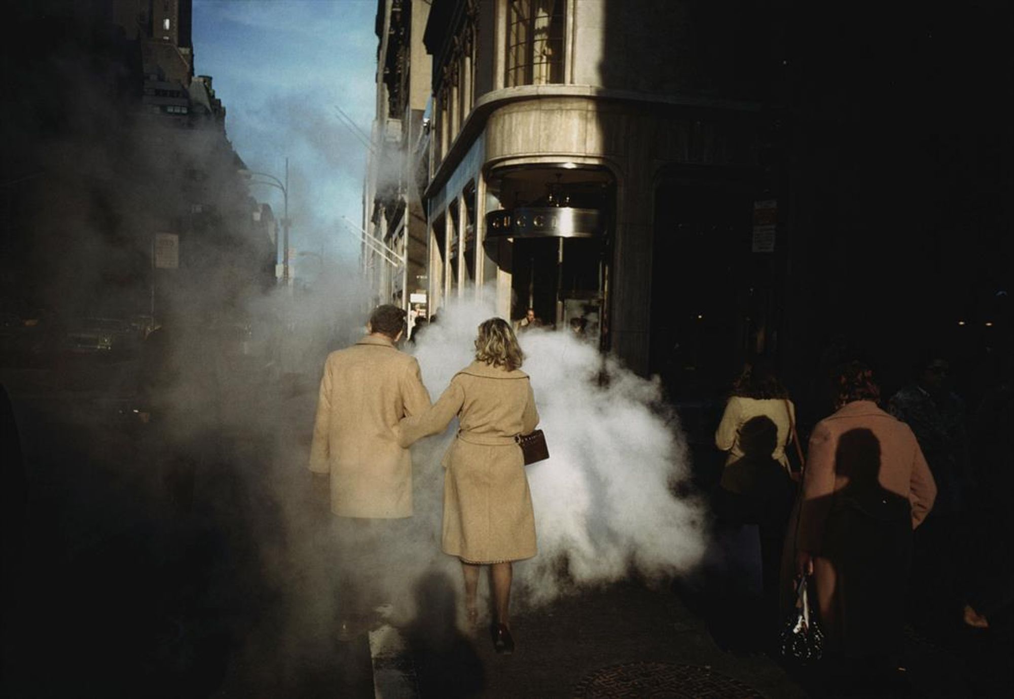 L'immagine raffigura una scena di strada dalla forte atmosfera urbana, catturata nei toni dell'ombra e della luce. Due persone, un uomo e una donna, si allontanano dalla telecamera attraverso una nuvola di vapore su un marciapiede cittadino. Entrambi indossano cappotti beige, mentre la donna porta con sé una piccola borsetta scura. Sembra che il vapore provenga da una grata sulla strada. La scena è prevalentemente in ombra, con un raggio di sole che illumina parte dell'edificio sulla destra, mettendone in risalto l'ingresso. Sullo sfondo sono visibili diversi altri pedoni, parzialmente oscurati dal vapore e dalle ombre circostanti. Il paesaggio urbano comprende edifici alti e un cielo azzurro sopra. 