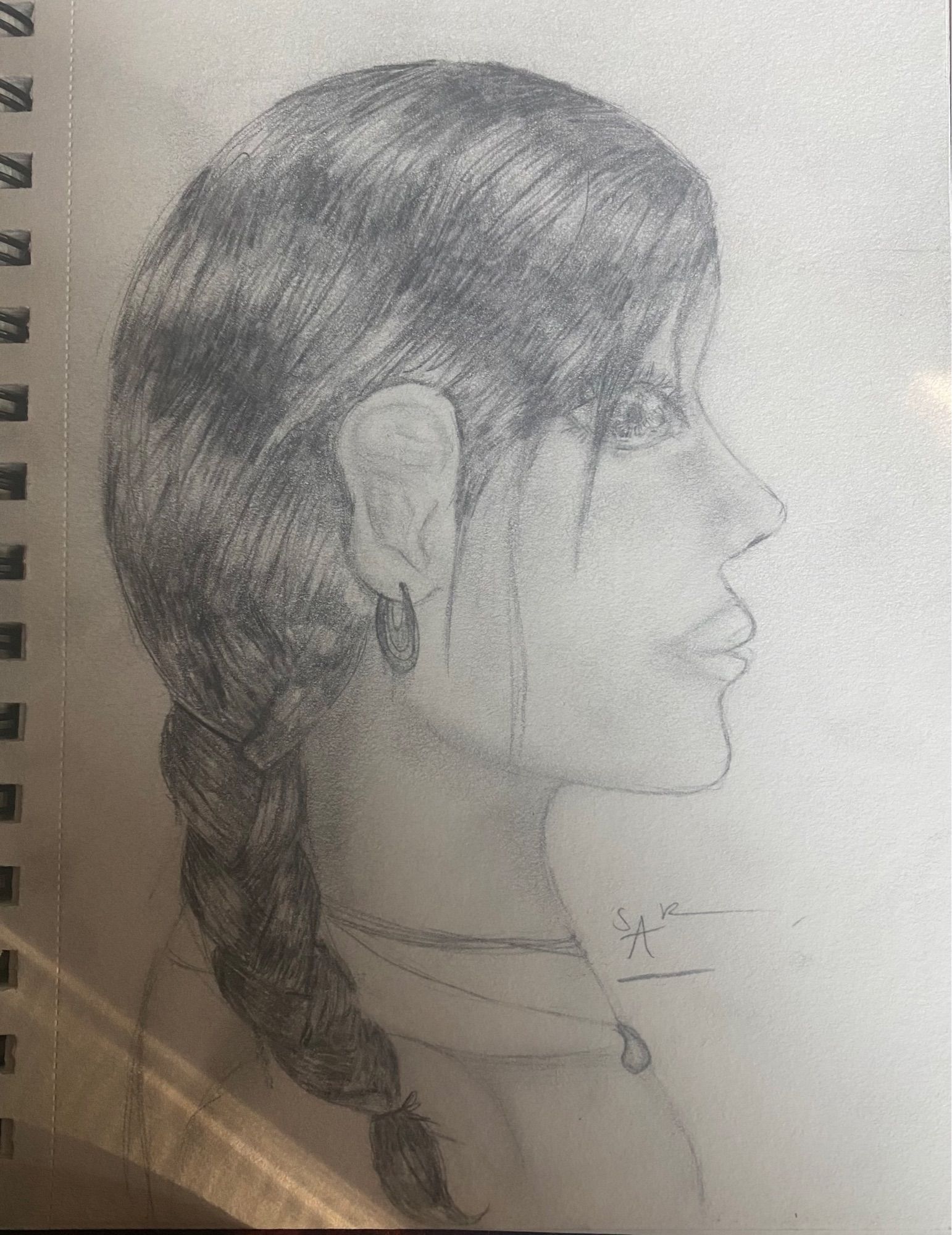 Pencil sketch in a notebook of a young woman looking right in profile. Her hair comes down over her shoulder in a braid. She has a small teardrop necklace and a little looped earring.