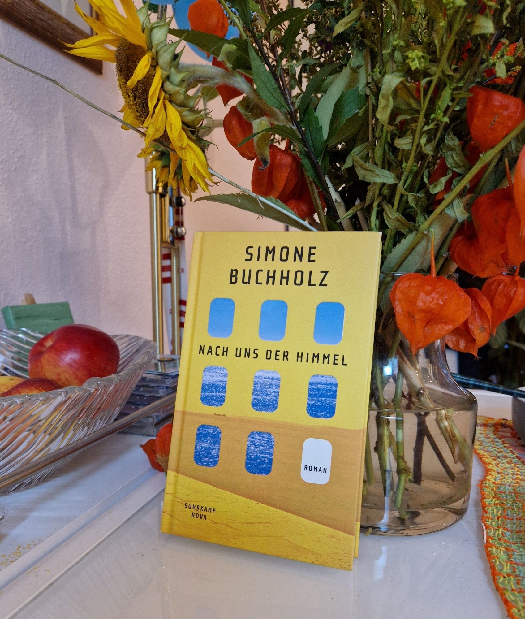 Buch: Simone Buchholz: "Nach uns der Himmel". Arrangiert mit Vase, Herbst Lumen, Obstschale, Tischdecke.