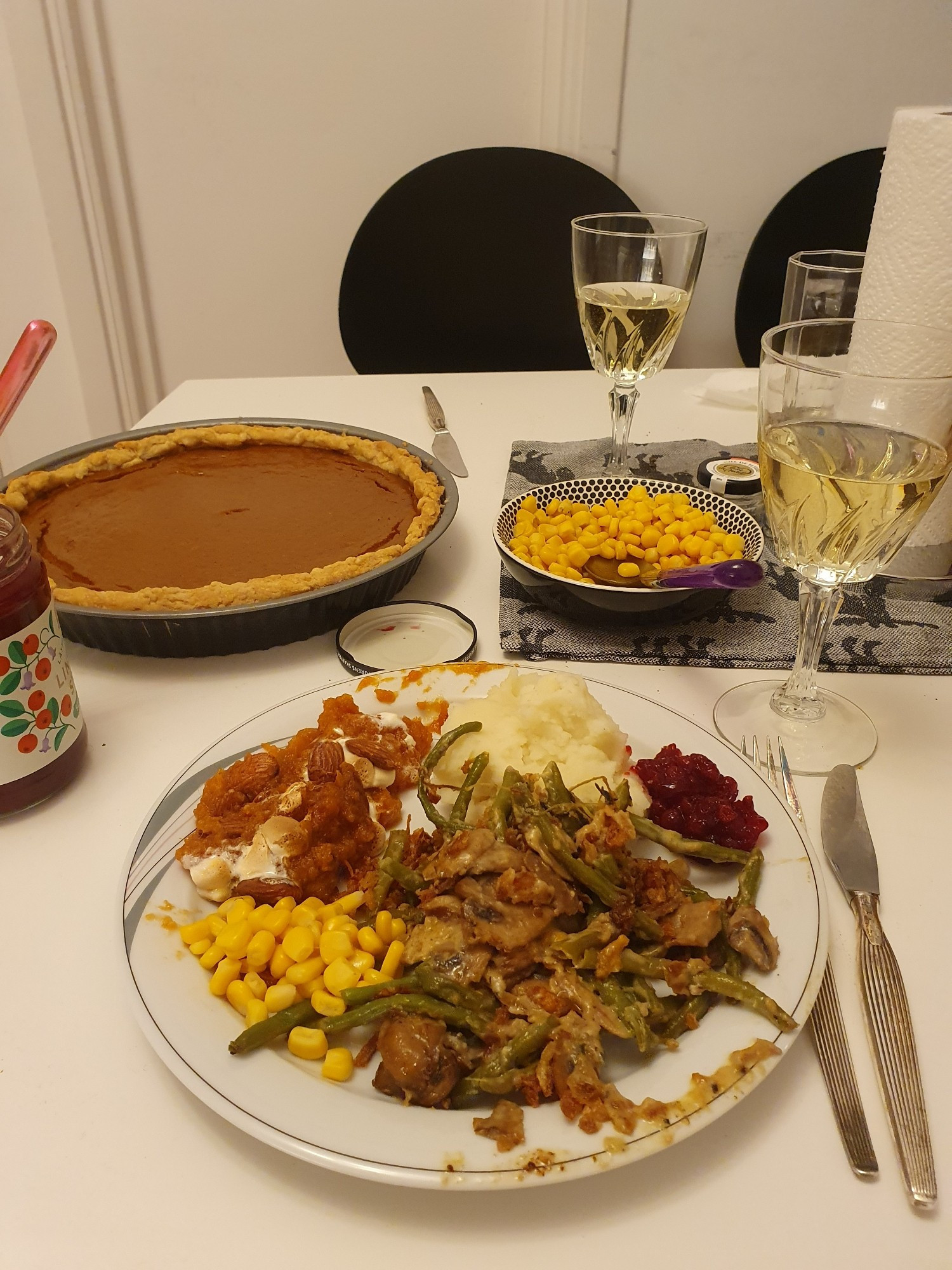 A plate of various thanksgiving dishes, like green bean casserole, mashed potatoes and sweet potato casserole. There's also two glasses of white wine and a pumpkin pie.