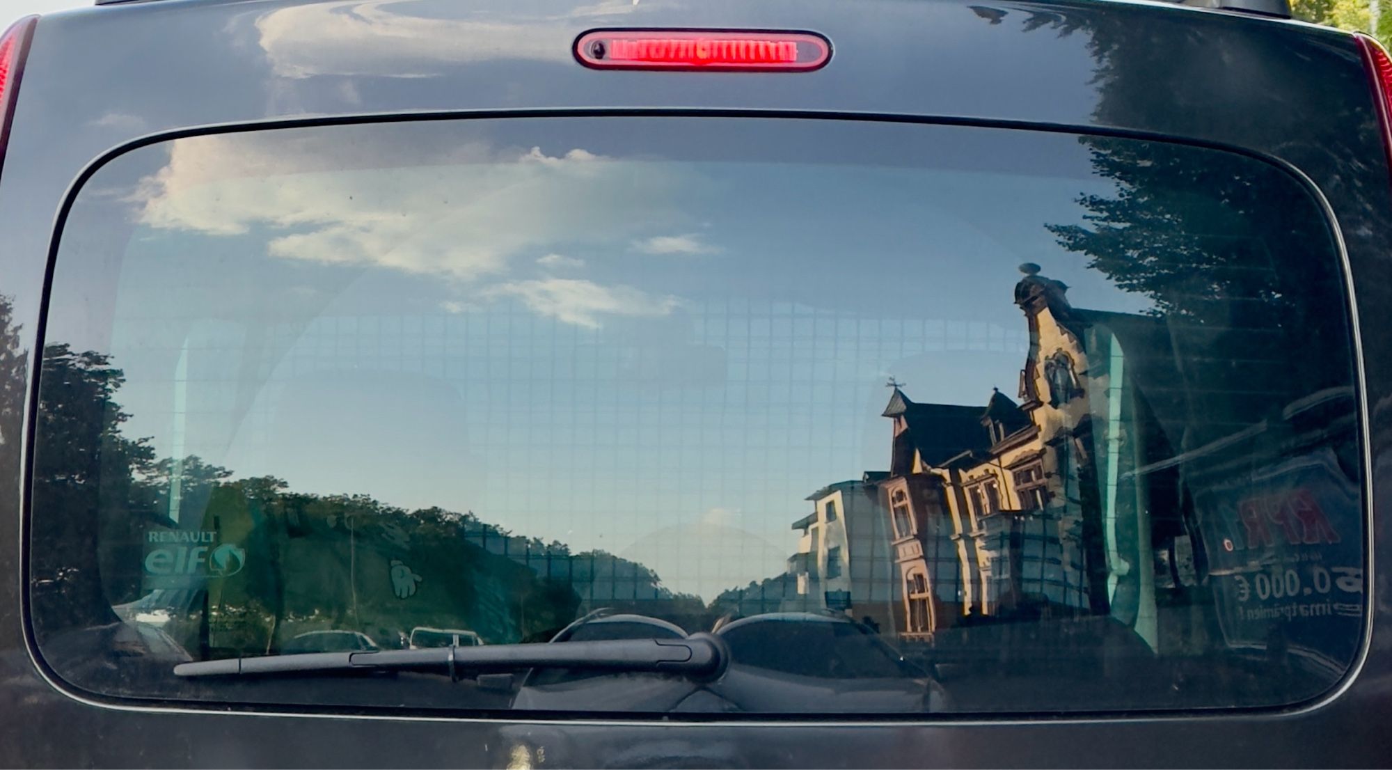 Eine Straßenansicht, Häuser, gespiegelt in einer mit Folie abgedunkelten Heckschreibe des vorausfahrenden Autos.