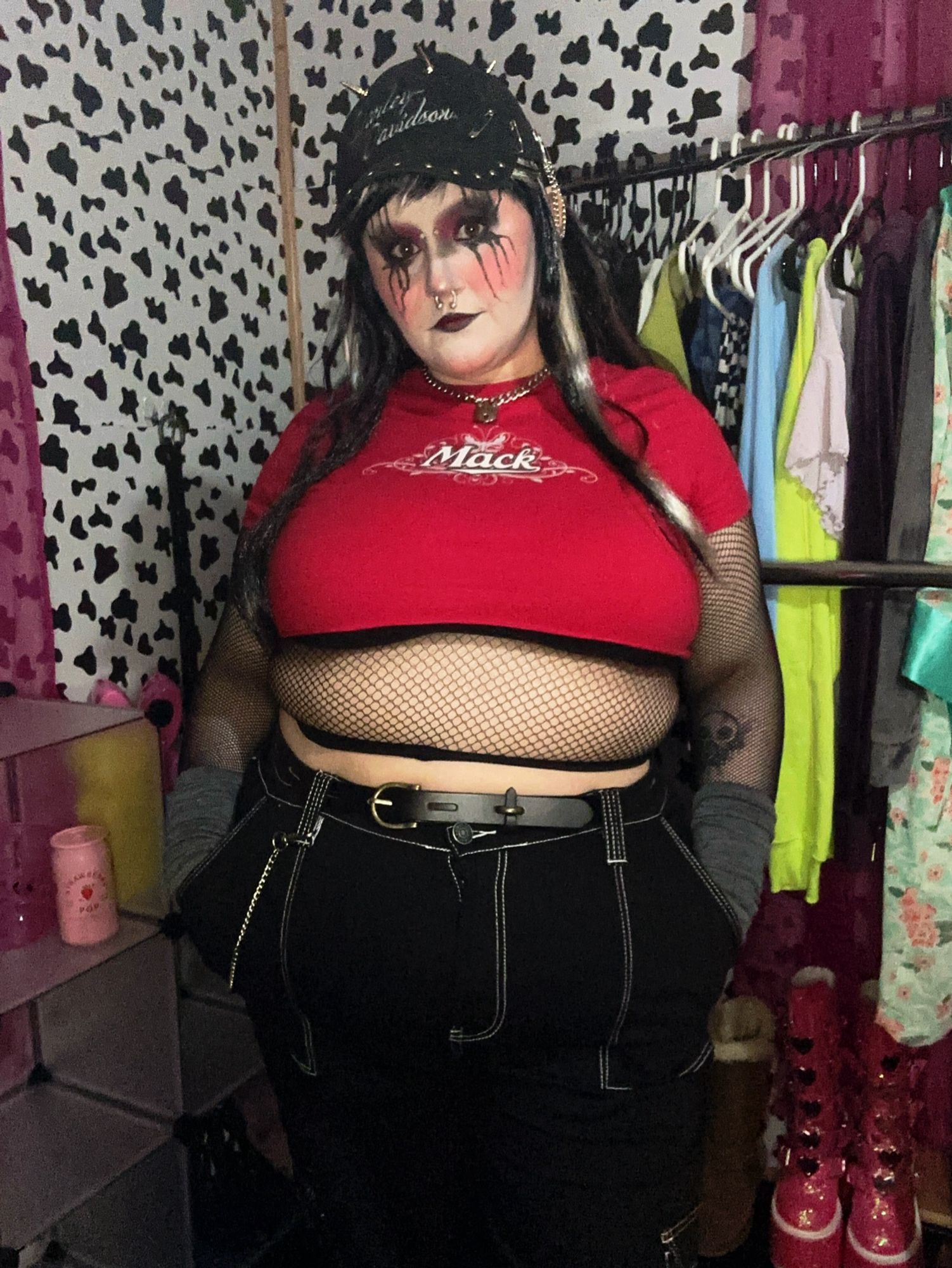 Page stands in front of a cow print backdrop and a clothing rack. They are dressed up in a “nu metal” or “punk” kind of attire. They have on: a black hat with spikes and chains, a red crop top with a black bra peeking out, a mesh top that covers their belly and arms, grey arm warmers, black cargo pants, and a skinny belt. Their face is painted white with red blush, black and red eyeshadow, messy black tendrils coming out from the eyeshadow, and red and black lipstick. They are also wearing a grey and black stripped costume wig.