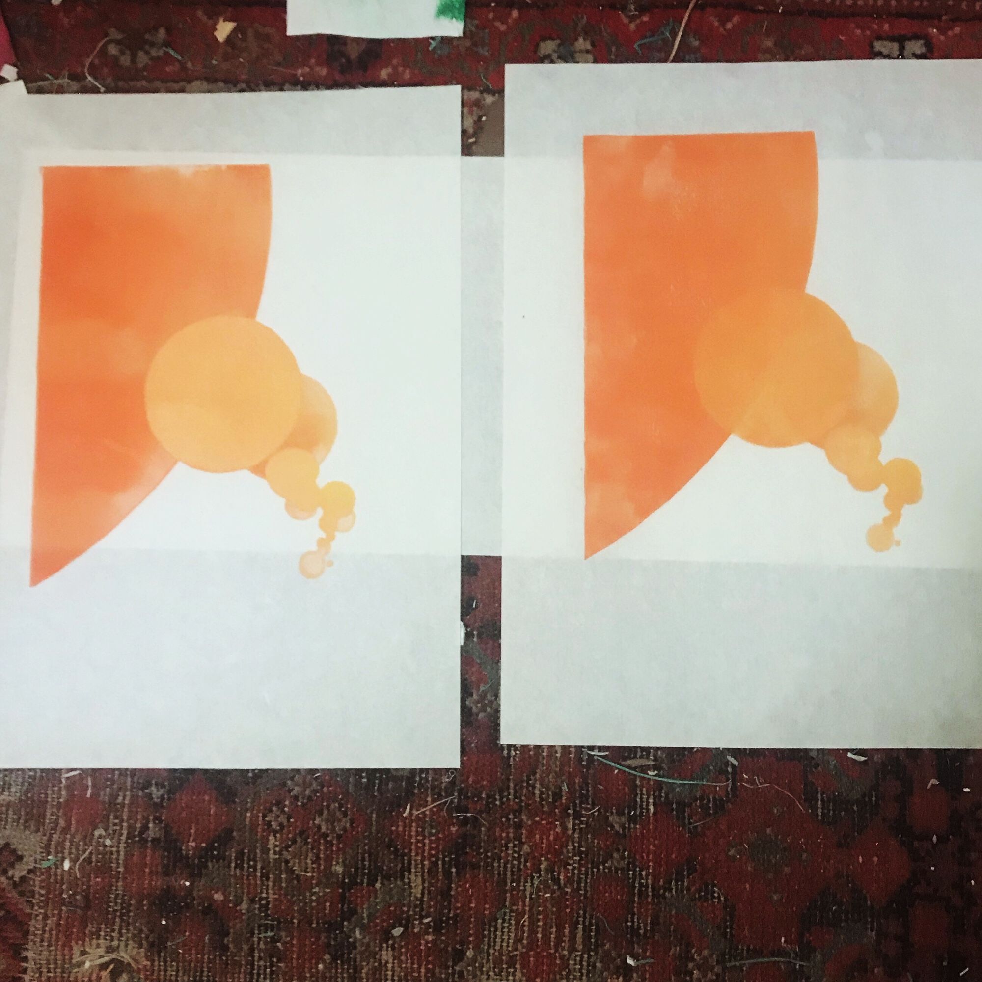 Two prints drying on the floor carpet. Each print is on white kozo paper, 30 cm by 40 cm and is printed in a gradient of orange to paler yellow-orange. The prints show a large section of a circle on the upper left through a smattering of smaller circles of various sizes trending to the lower right. Now it its second stage the prints show overlapping circles.