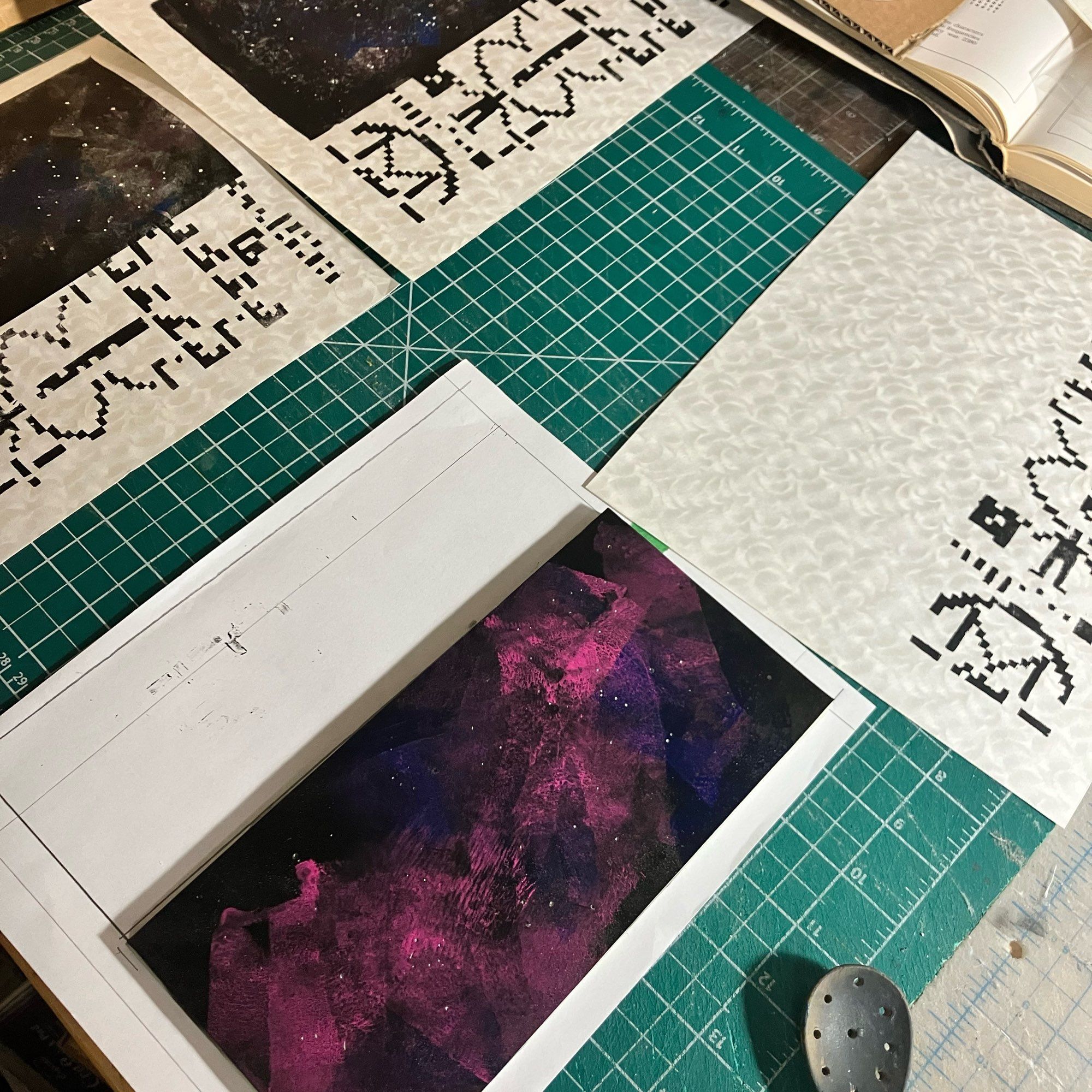 A table top with cutting mats and prints in various stages: there are two completed prints on white Japanese Obonai paper with a feathery sheen with the Arecibo Message printed in black like a pattern of pixels with some easily identifiable shapes like a human, satellite dish and double helix in a vertical rectangle along the right side with a vertical rectangle on the left as a starry space image with some swirling pinks and blues along with black. There is a third print only printed with the Arecibo Message. There is an inked plate for the space rectangle ready to print on top of a paper alignment jig. You can also see a roll of tape, open book and wooden spoon at the edges of the photo.