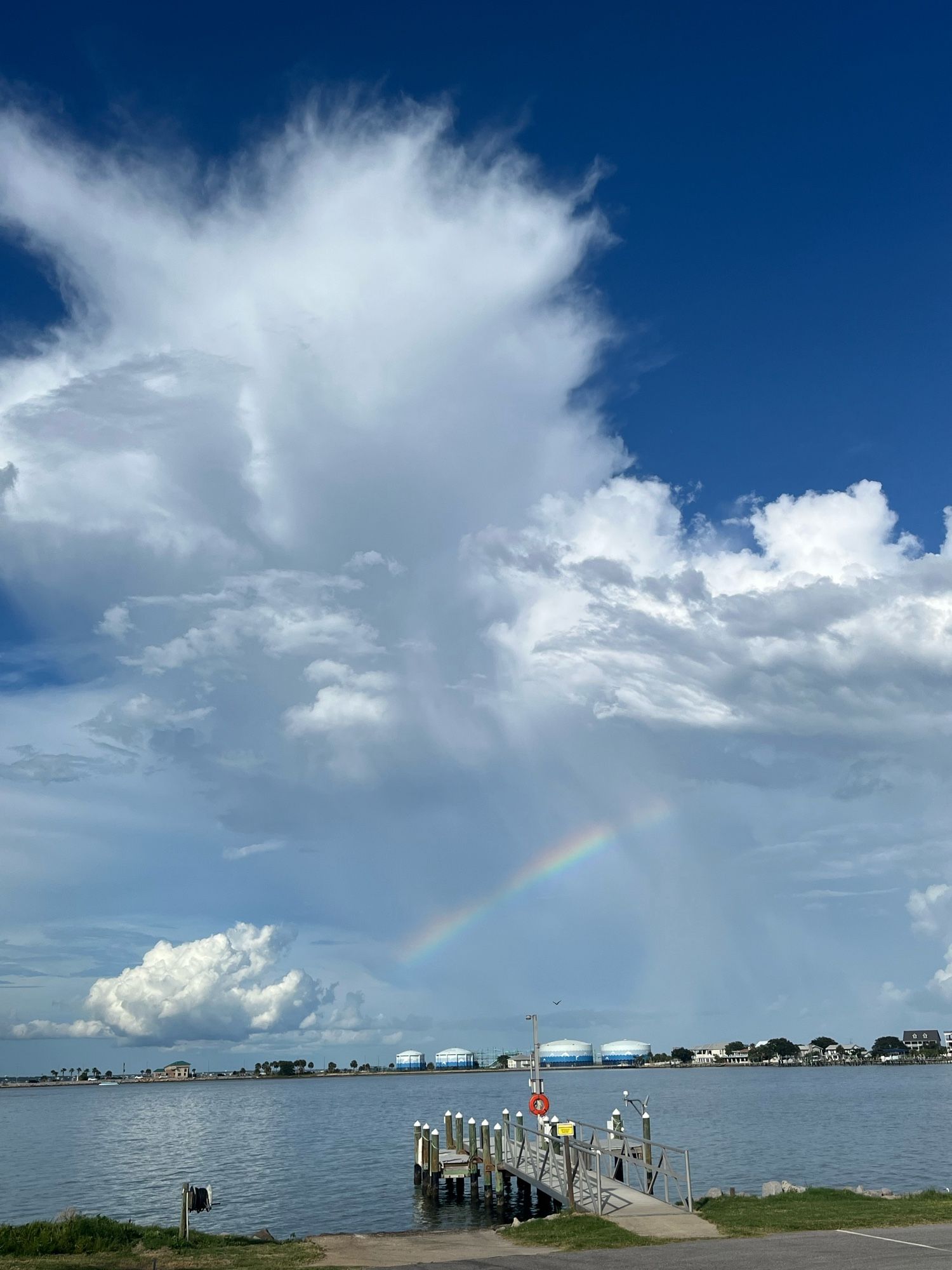 White clouds in front of a blue sky, and half a rainbow underneath, over a body of water and a dock