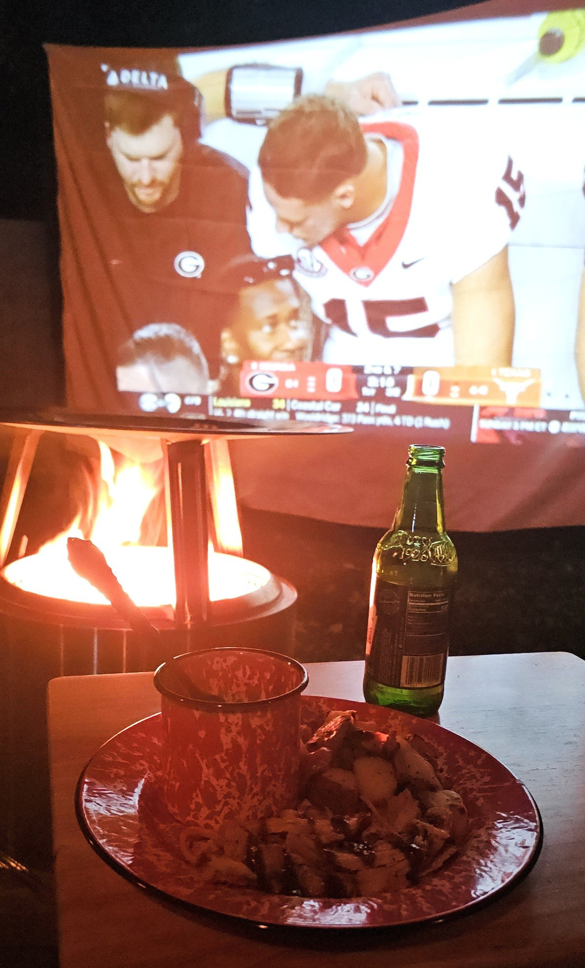 A tray of food in the foreground with an outdoor fire and a projected screen of football in the background 