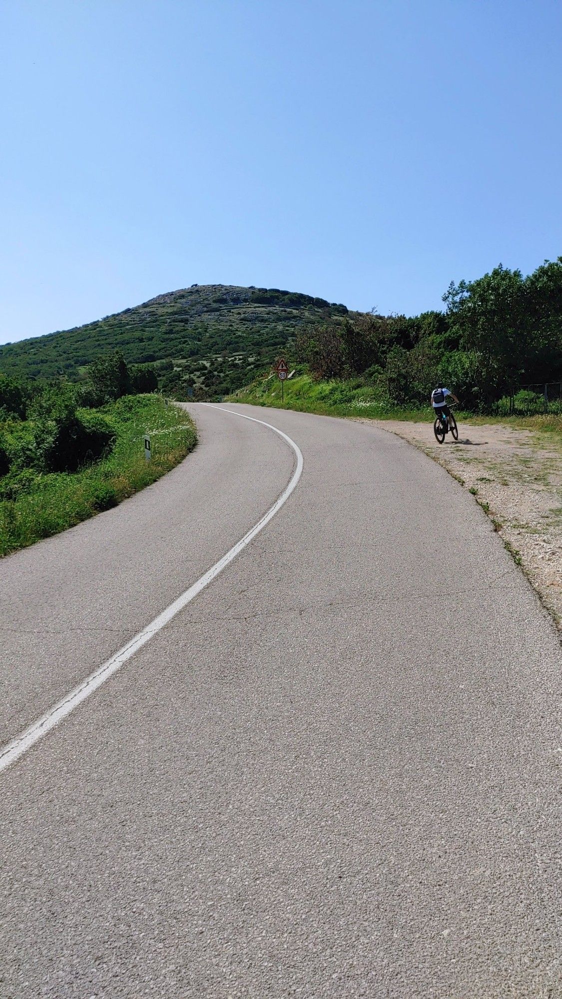 Leichte Linkskurve mit durchgehendem Mittelstreifen, Blick auf grünen Hügel, rechts am Rand steht ein Fahrradfahrer