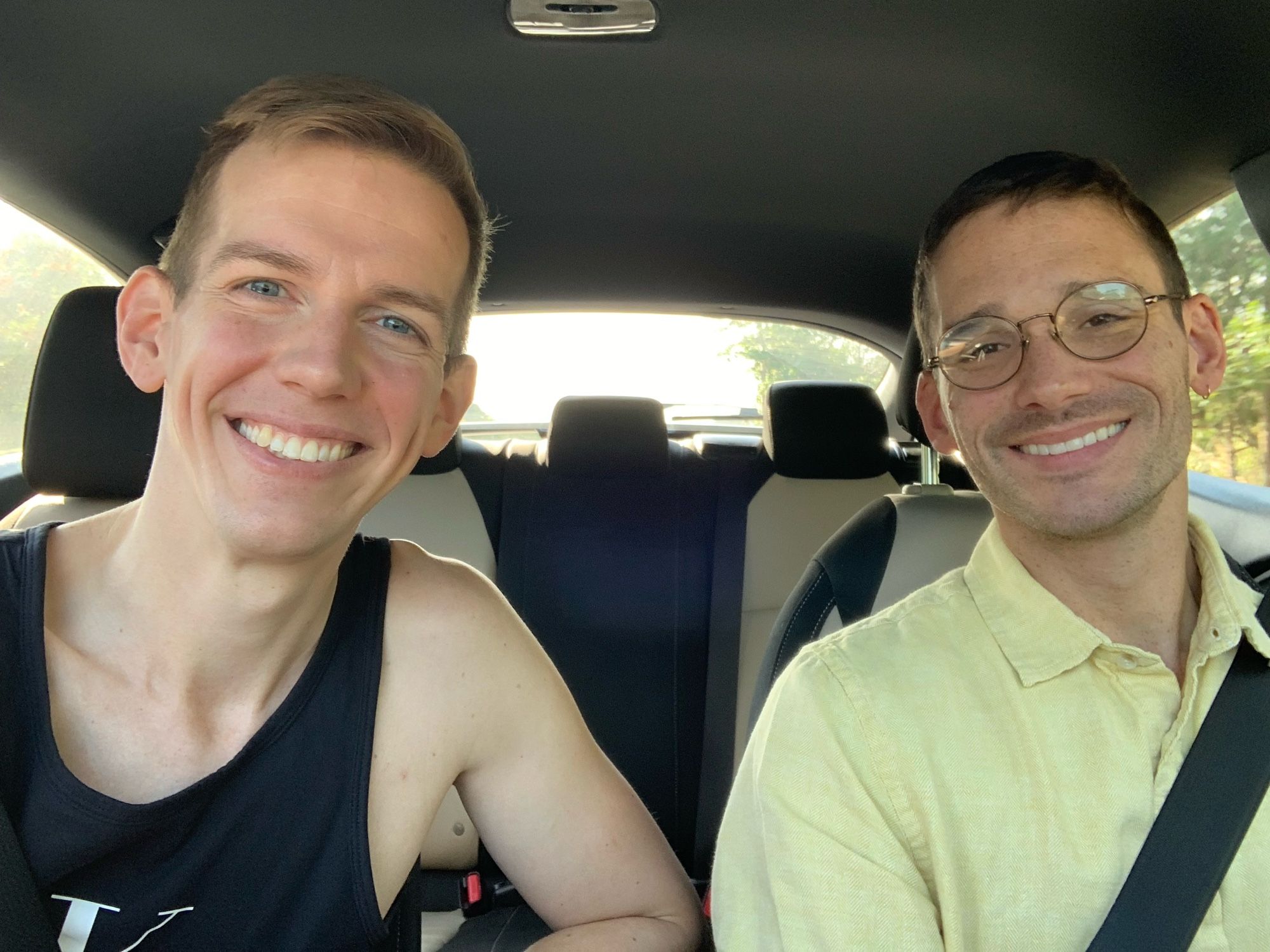 Two gays sitting in their moving vehicle, smiling, right before they discovered that they forgot their beach towels and sunscreen at home, so they had to find a local beach boutique to purchase them at an unbelievable price 🫠