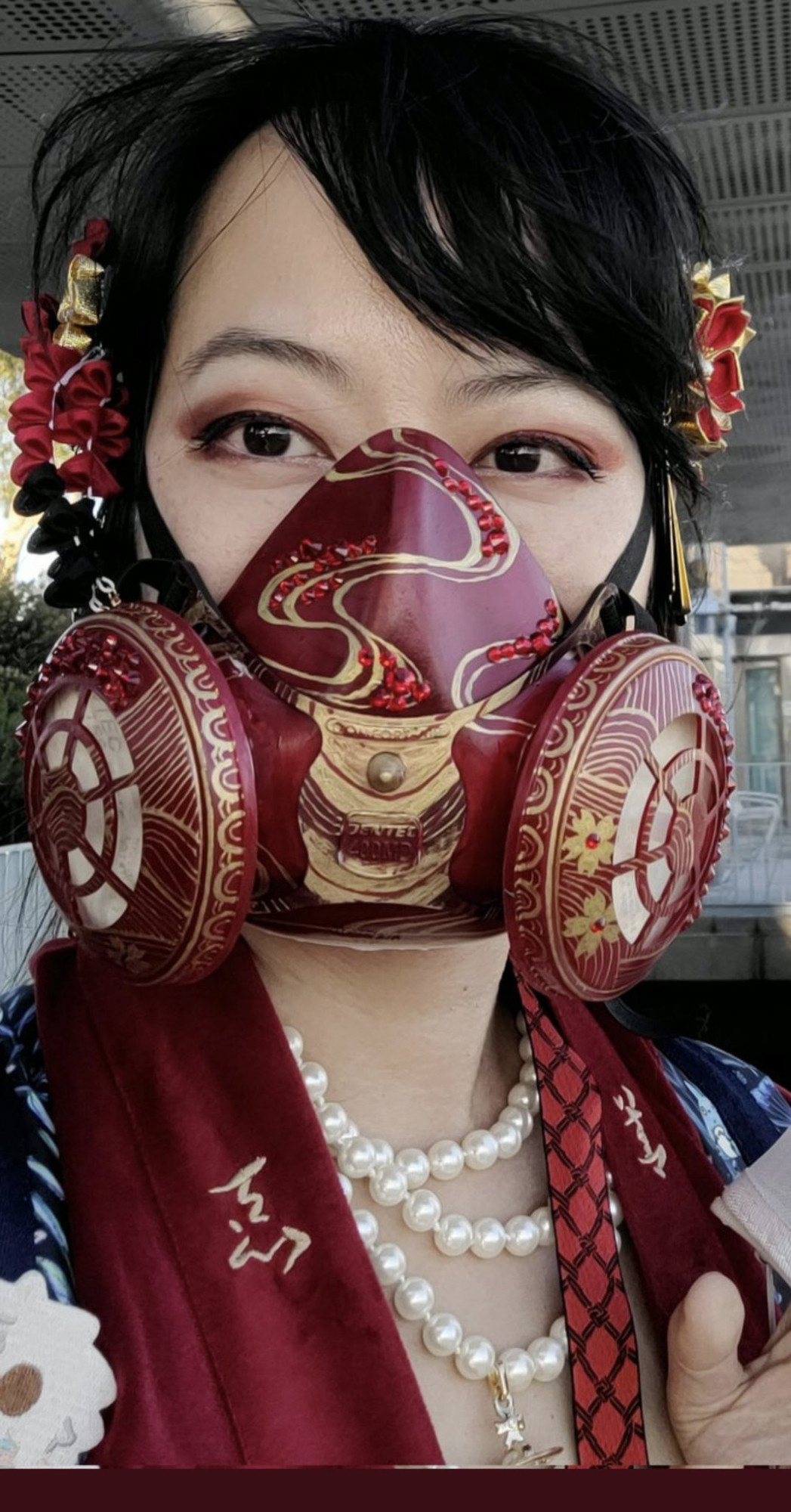 color photo of femme person wearing an elastomeric mask hand-painted in red and gold. The design takes inspiration from historical Japanese lacquerware design. The facepiece features flowing water patterns accented by red crystal embellishments. The filter covers are accented with line art of mountains with a spread of red crystals across the top, cherry blossoms. and an outer border of seigaiha waves along the curve.