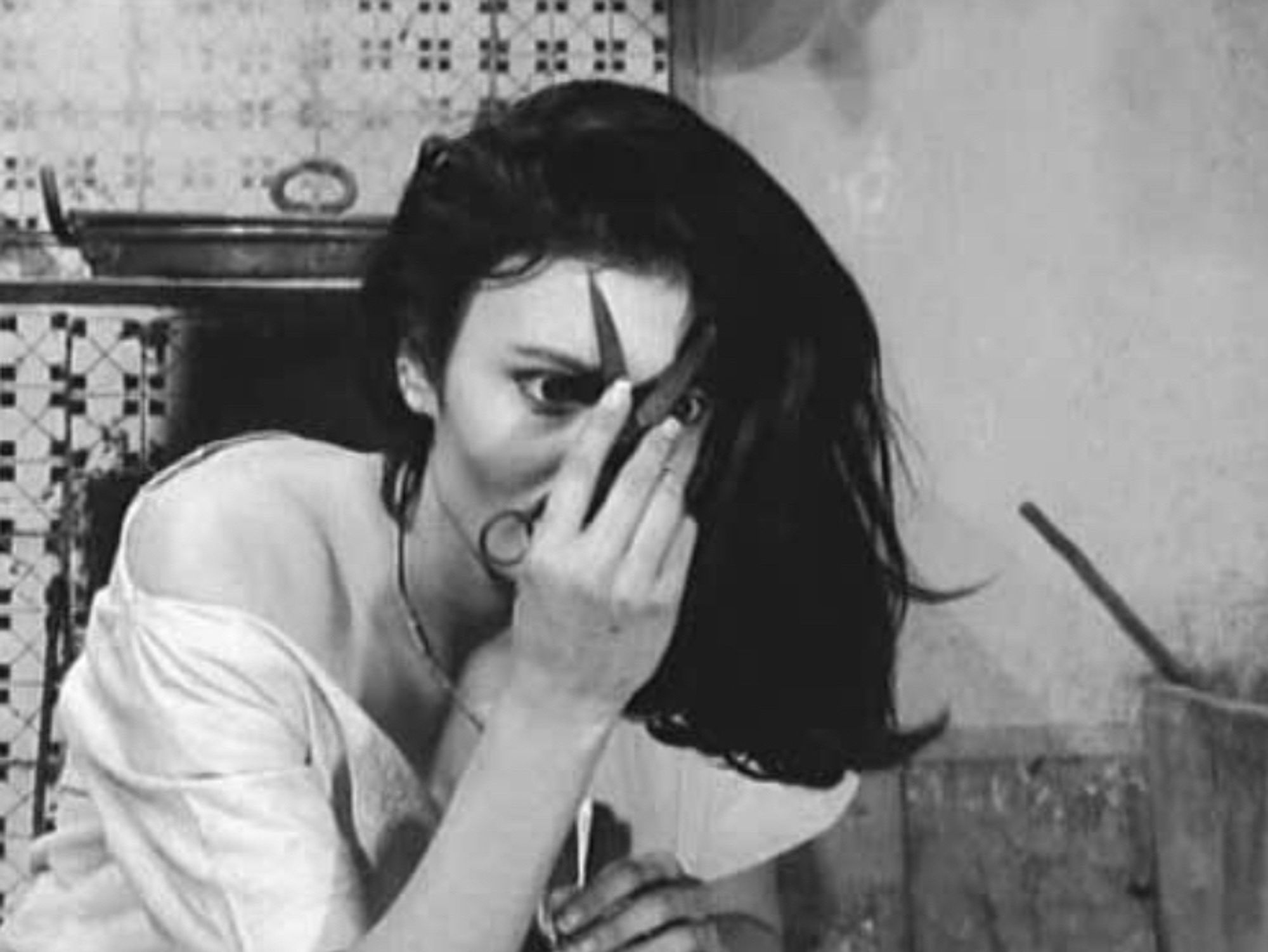 A black and white still image of a beautiful but disheveled young woman with raven black hair. She is holding an open pair of scissors flat against her forehead, which is very unsettling. The scissors give the impression that she has horns.