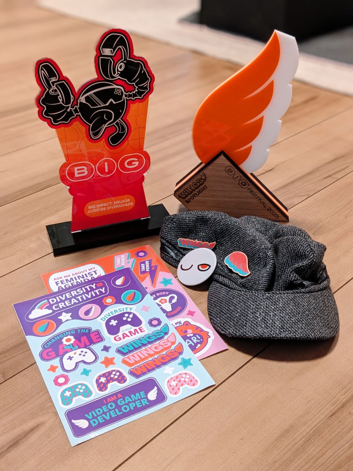 a "BIG: Big Impact" trophy with a robot character in it and a "WINGS Spotlight" trophy with a wing on it behind two sets of stickers featuring gaming and diversity and a cap with three pins on it