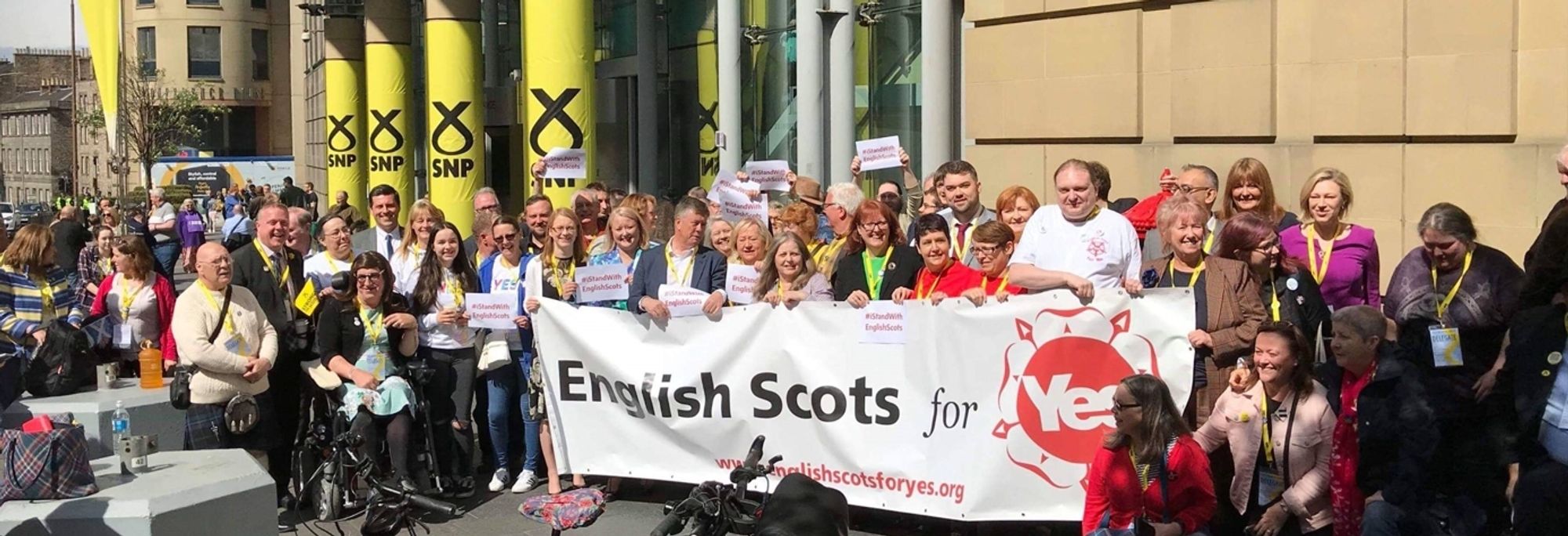 A crowd of smiling supporters of Scottish independence outside an SNP conference with a banner for the group English Scots for Yes