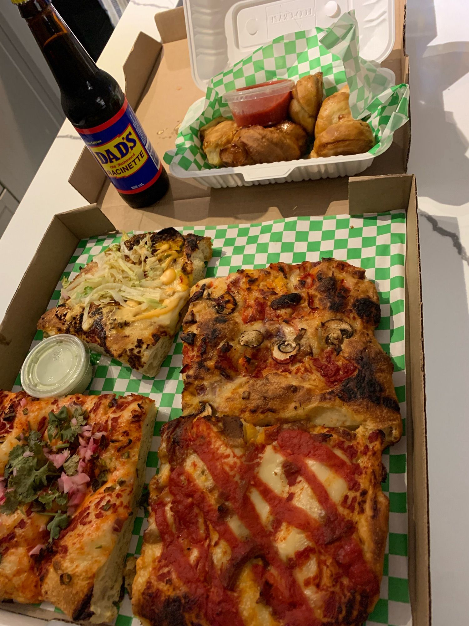 Photo of 4 big square pizza slices, all with different toppings. They sit in a pizza box with green checkered paper. Just being them is a box of garlic knots