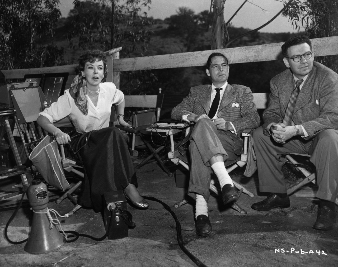 Lupino leaning forward in director’s chair, megaphone and amplifier on ground just in front of her. Young and Wald sit in their own director’s chairs next to her. Natural hilly scene behind them.