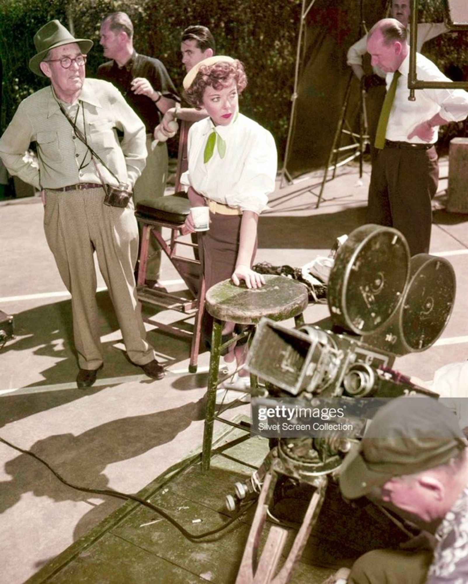 Color photo of Director Lupino and crew outdoors; Lupino surveys scene while cameras are set up. Cinematographer Archie Stout stands near Lupino, wearing a hat. Assistant Director James Anderson in dark shirt in the background.