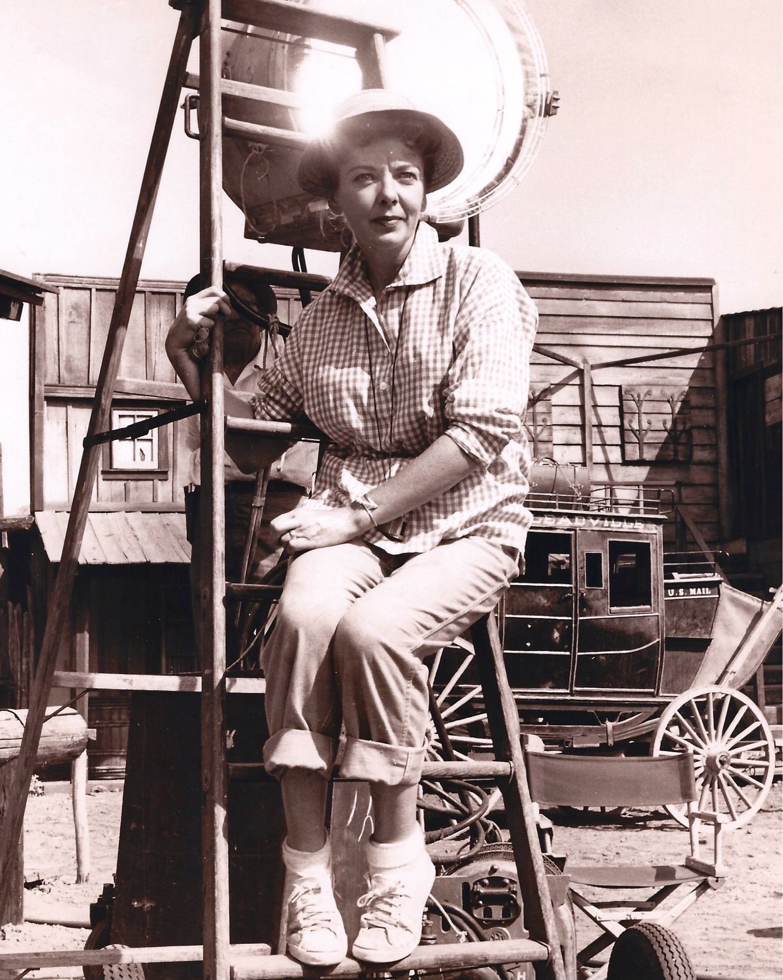 Lupino, wearing casual clothes, sits on a ladder amidst an outdoor set of an Old Western street