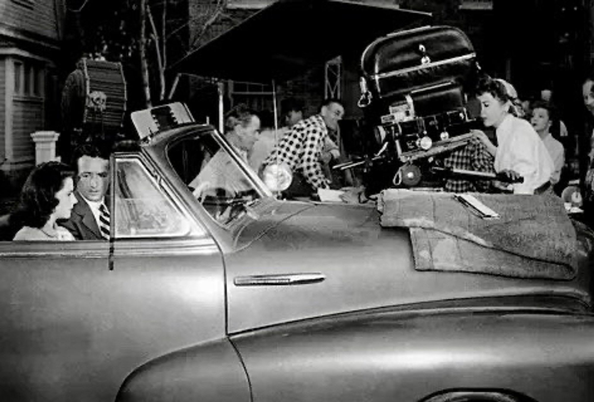 Powers and Clarke sitting in a open-roofed car. Lupino eyes scene through large movie camera pointed at them. Various crew members in background.