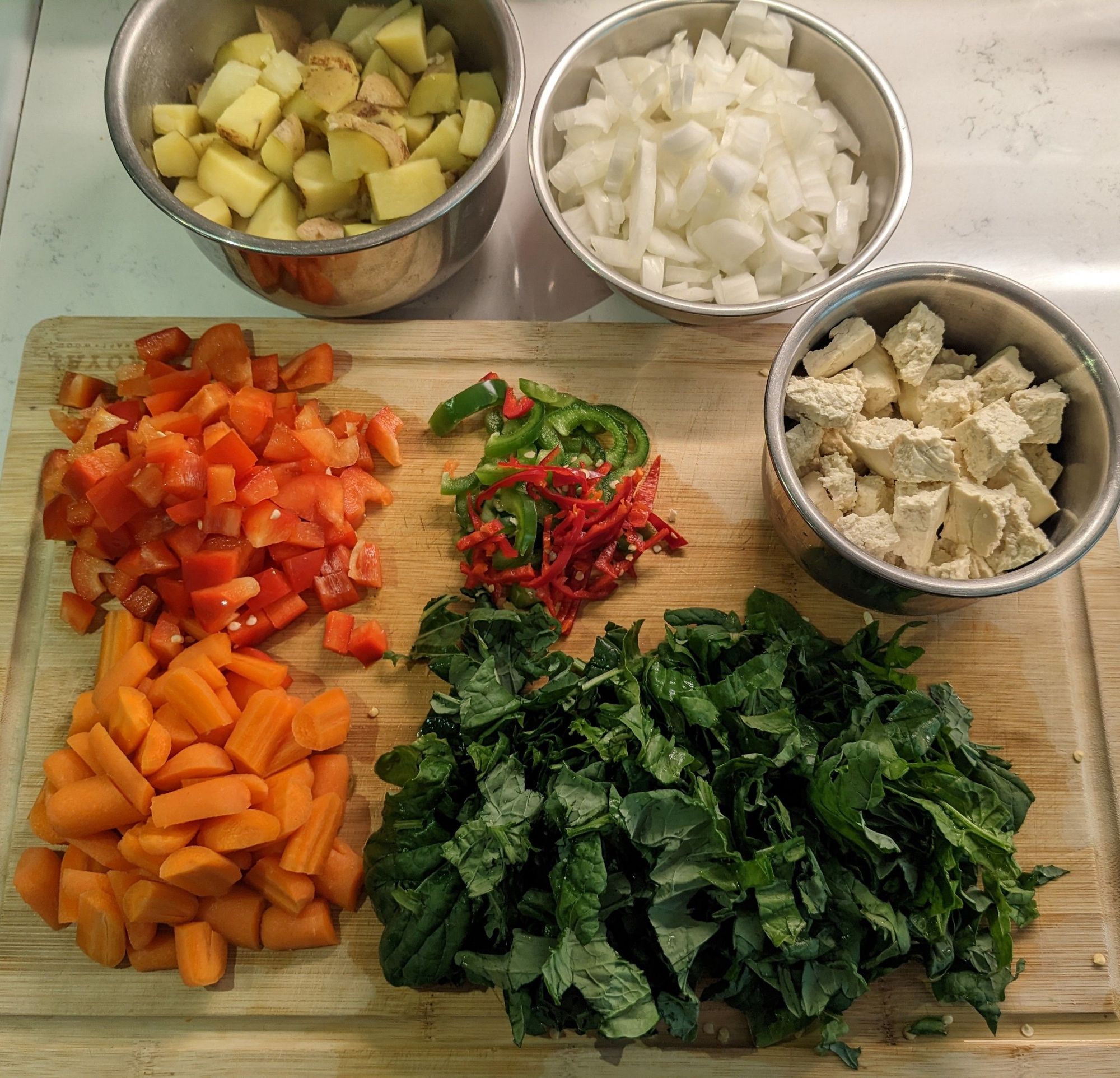 An arrangement of raw and cooked chopped vegetables on a cutting board and in stainless steel bowls: steamed and chopped potatoes, raw onion, raw sweet peppers, raw hot peppers, raw carrots, raw spinach and Chijimisai, hand-crumbled tofu.